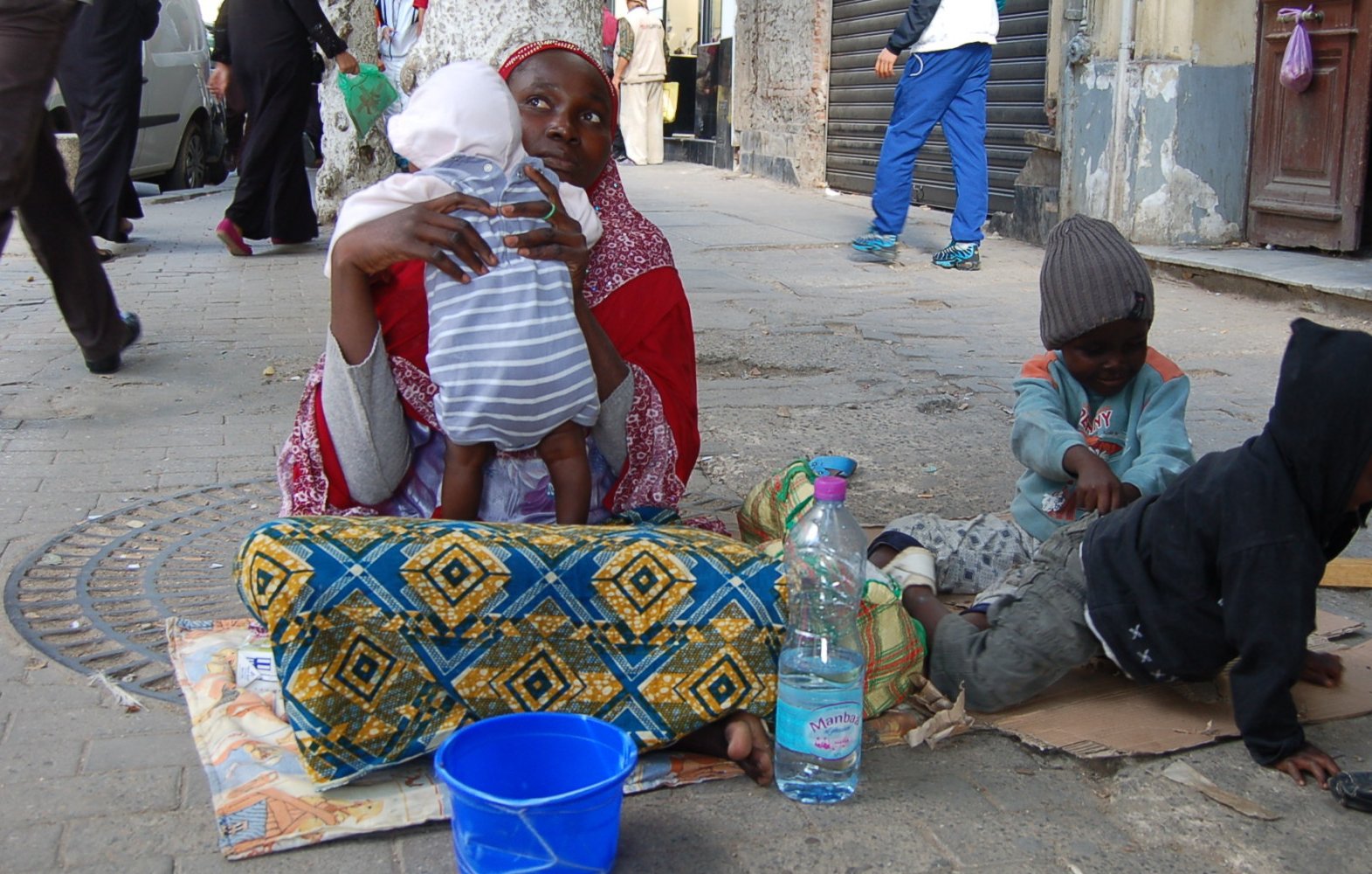 L'image montre une femme assise sur le sol, tenant un bébé dans ses bras. Elle est entourée de deux enfants, l'un assis à côté d'elle et l'autre jouant sur le sol. La femme porte un vêtement coloré et un foulard, tandis que les enfants semblent habillés de manière simple et modeste. À côté d'eux, il y a une bouteille d'eau et un seau bleu. La scène se déroule dans une rue, avec des bâtiments en arrière-plan et d'autres personnes qui passent. L'ambiance semble à la fois familiale et difficile, évoquant des thèmes de pauvreté et de lutte quotidienne.