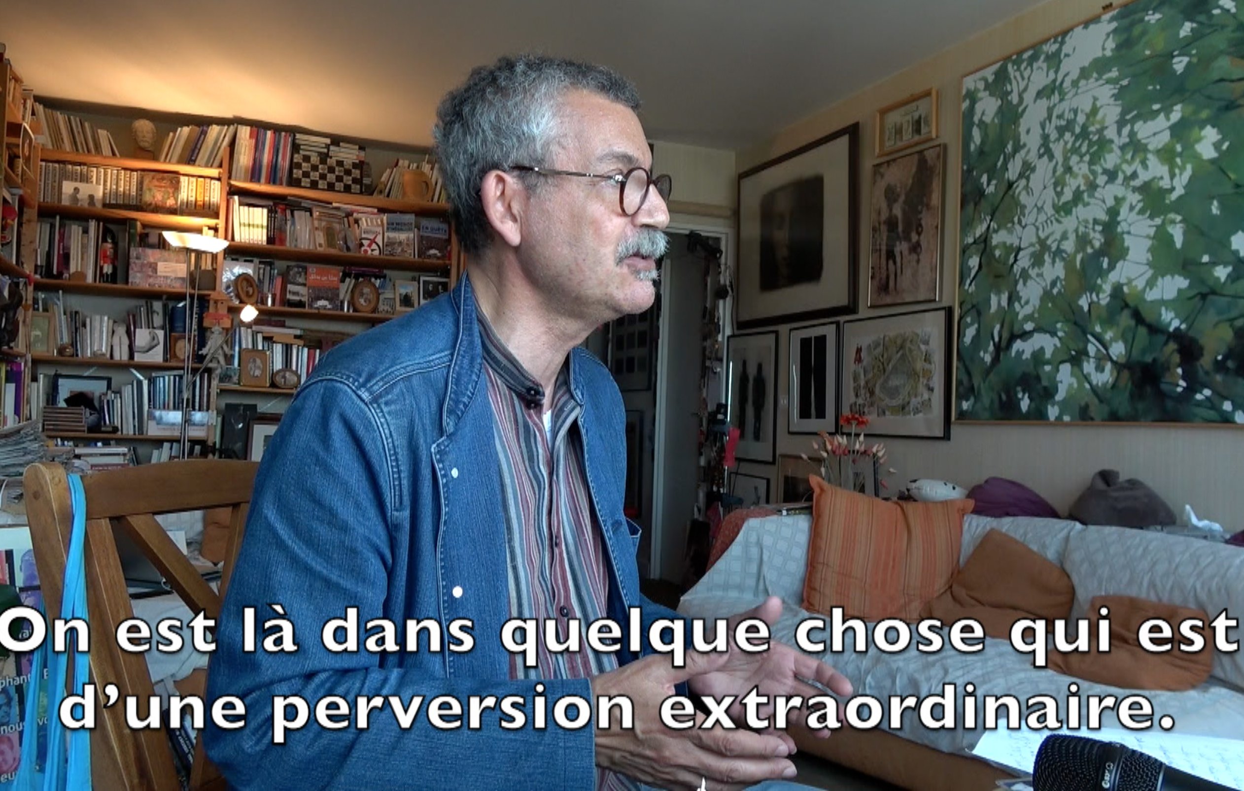 L'image montre un homme assis à une table, parlant avec animation. Il semble s'exprimer sur un sujet profond, probablement lié à des thèmes intellectuels ou sociaux. En arrière-plan, on peut voir des étagères remplies de livres et plusieurs œuvres d'art accrochées aux murs, ce qui indique un environnement artistique et studieux. Le texte sur l'image suggère qu'il aborde des concepts de manière critique, impliquant une réflexion sur des questions de moralité ou de société.