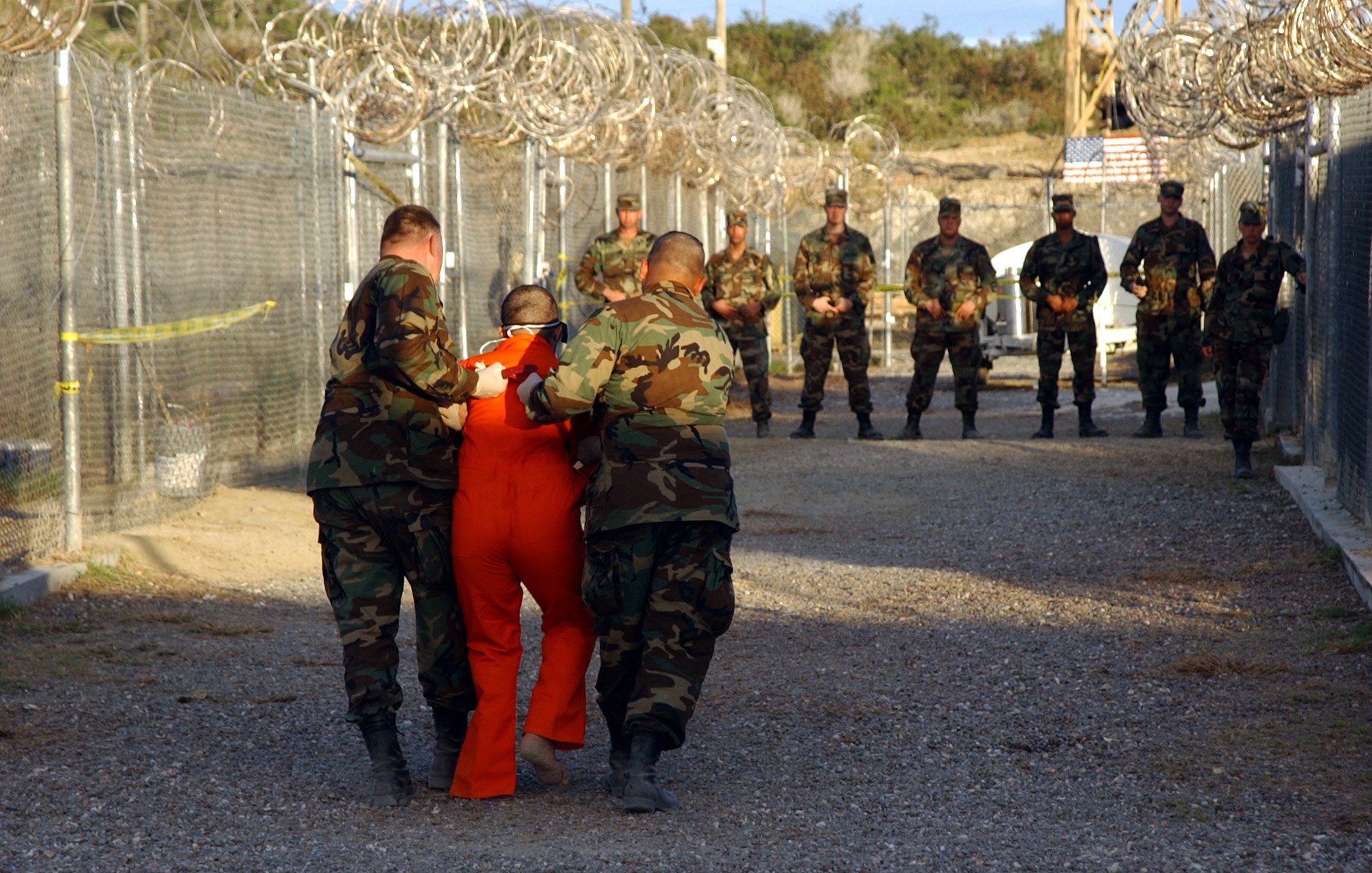 در این تصویر، چند نظامی در لباس‌های نظامی در یک فضای محصور با دیوارهای مشبک و نوارهای خاردار حضور دارند. یکی از نظامیان در حال هدایت یک فرد در لباس نارنجی است که به نظر می‌رسد دستگیر شده باشد. تعدادی دیگر از نظامیان در پس‌زمینه ایستاده‌اند و به صحنه نظاره می‌کنند. فضا به شکل یک محوطه امنیتی یا زندان به نظر می‌رسد.