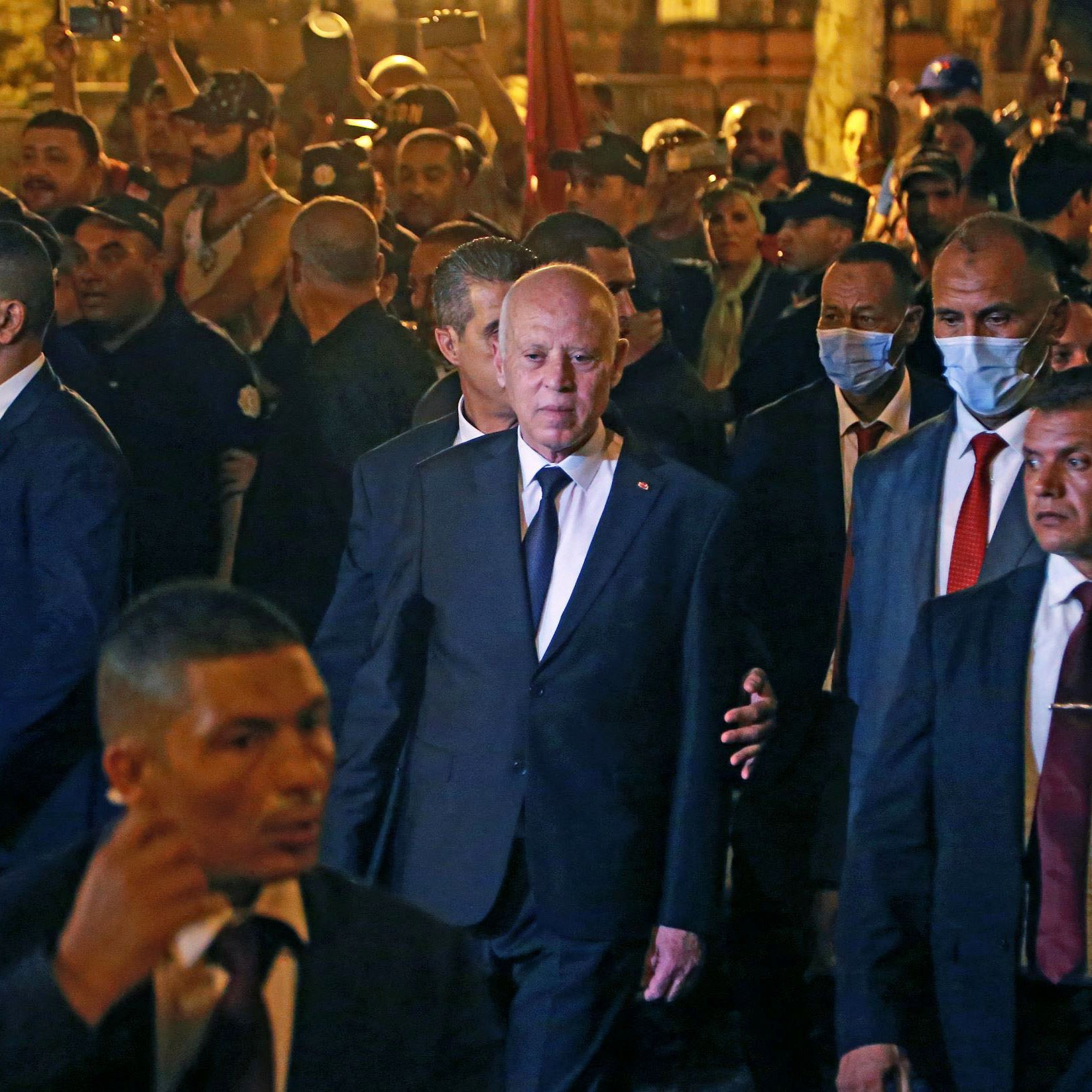 L'image montre un groupe de personnes rassemblées autour d'un homme en costume noir, qui semble être une figure politique ou publique. Il est entouré de plusieurs membres de ce qui semble être une équipe de sécurité, tous vêtus de costumes sombres. Les gens en arrière-plan semblent attentifs et certains utilisent des téléphones pour prendre des photos. L'ambiance paraît animée, avec un éclairage qui suggère qu'il fait sombre.