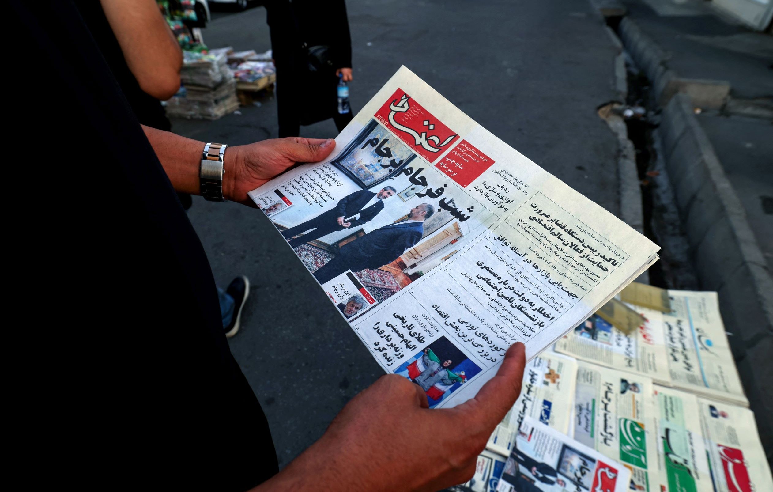 L'image montre une personne tenant un journal ouvert. Le titre est bien visible et semble relater des informations d'actualité, potentiellement politiques ou sociales. En arrière-plan, on aperçoit un kiosque à journaux avec d'autres exemplaires de journaux exposés. L'environnement semble urbain, avec des voitures garées et un sol en pavé.