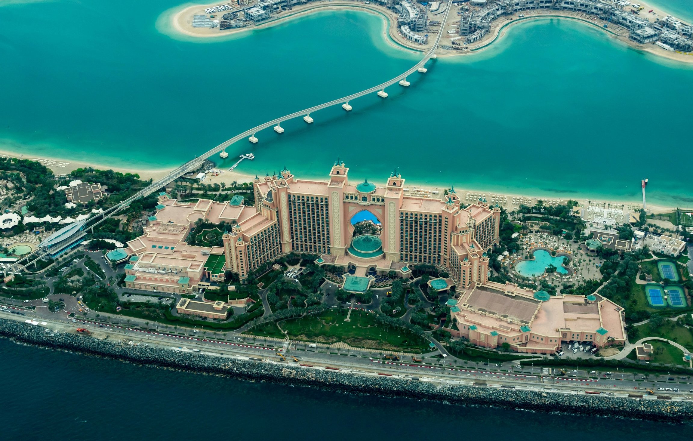 L'image montre une vue aérienne d'un impressionnant complexe hôtelier situé sur le bord de la mer. On peut voir un grand bâtiment aux couleurs beige et rose, entouré de jardins bien entretenus et de plusieurs piscines. La mer turquoise qui l'entoure contraste avec les structures urbaines modernes visibles en arrière-plan. Un pont relie le complexe à une autre zone, et l'ensemble est à la fois luxueux et captivant, évoquant une destination touristique prisée.