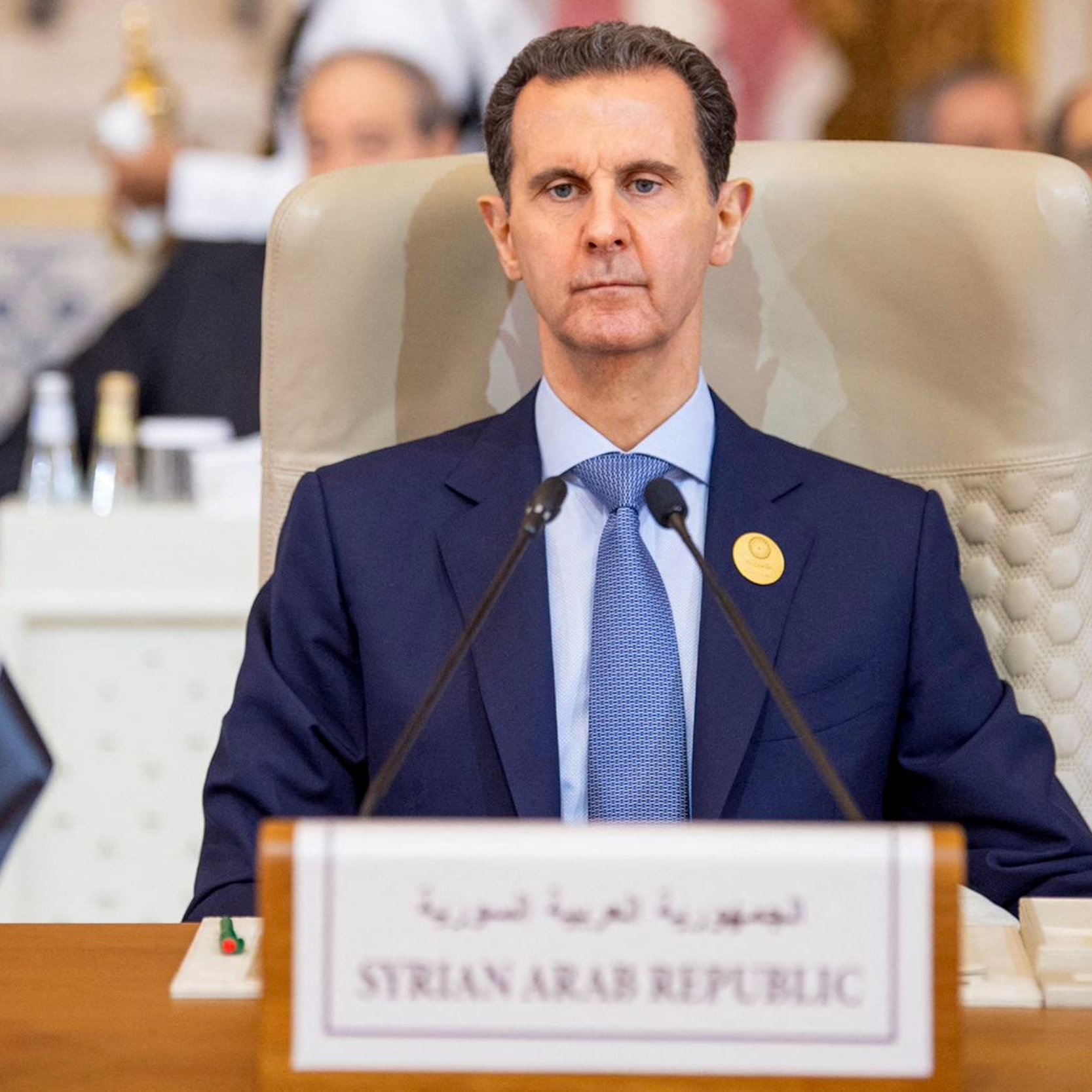 L'image montre un homme assis derrière un bureau lors d'une réunion officielle. Il est habillé en costume sombre et semble sérieux. À côté de lui, il y a un drapeau de la Syrie et une plaque indiquant "République arabe syrienne". En arrière-plan, on peut apercevoir d'autres personnes et une disposition formelle typique d'une conférence ou d'un sommet.