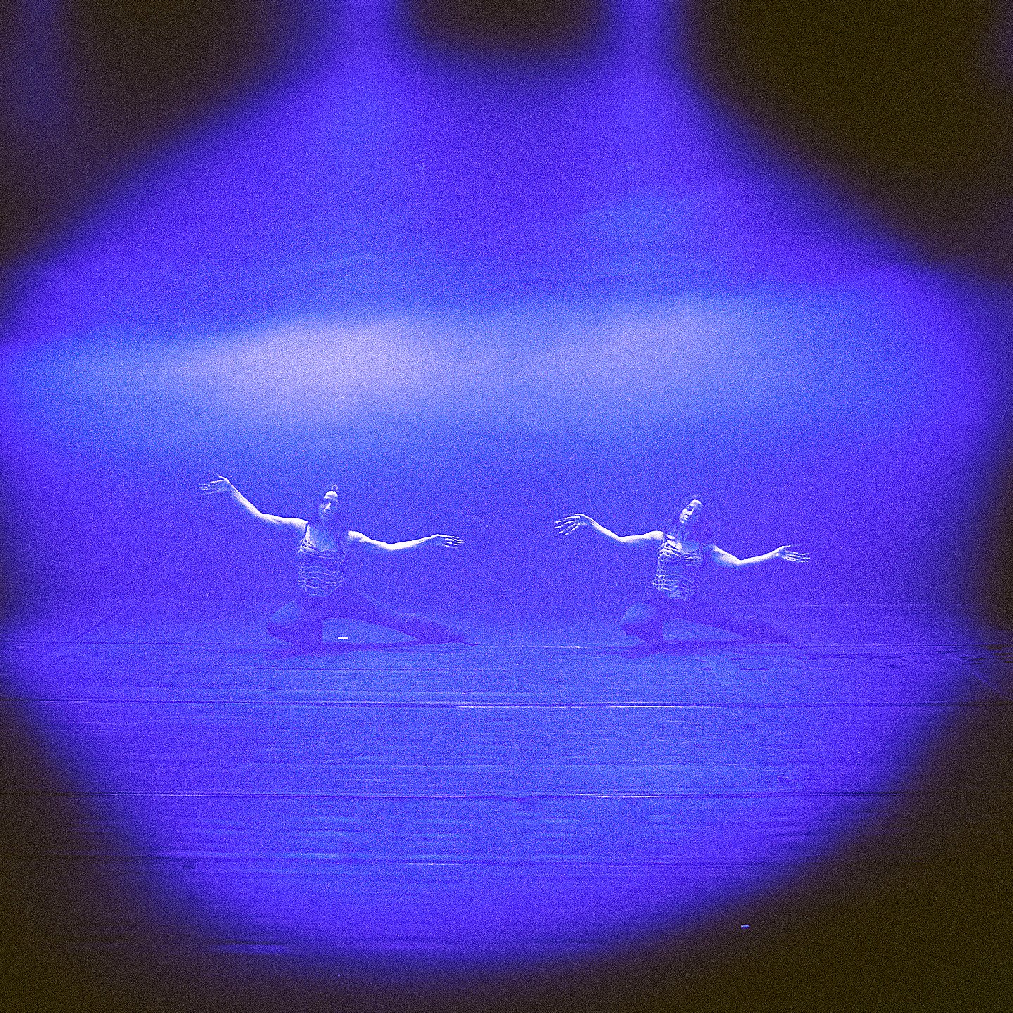The image features two dancers positioned on a stage, surrounded by a soft, dramatic purple light. They appear to be performing a synchronized dance, with their arms outstretched. The background is dark, drawing focus to the illuminated area where they are dancing. The scene conveys a sense of artistry and expression, emphasizing the dancers' movements and the emotional atmosphere created by the lighting.