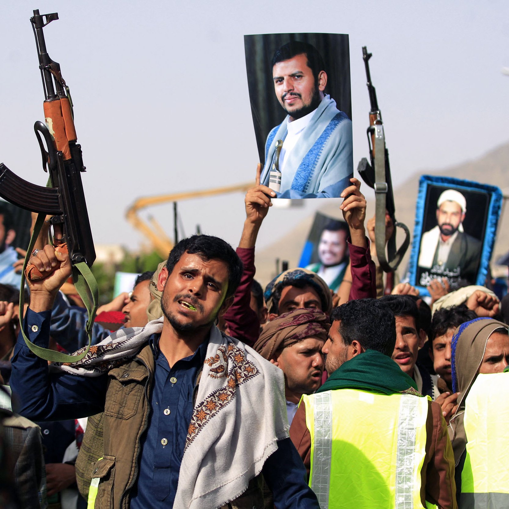 L'image montre une foule de manifestants tenant des fusils automatiques. Certains d'entre eux brandissent des portraits de figures emblématiques. Les gens semblent exprimer des émotions fortes, comme la détermination ou l'enthousiasme. L'environnement évoque une ambiance de rassemblement ou de manifestation politique. Des éléments comme des vêtements spécifiques et le paysage suggèrent un contexte culturel particulier.