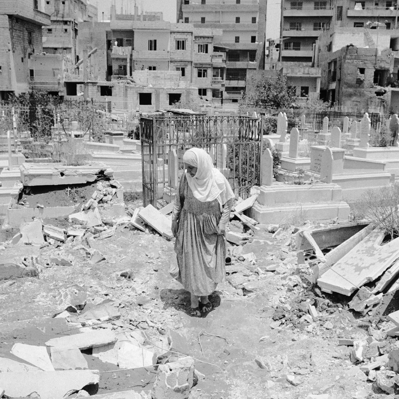 L'image montre une femme vêtue d'un long habit, marchant au milieu d'un cimetière. Le sol est désordonné, avec des débris éparpillés, ce qui suggère qu'il y a eu des destructions récentes dans la zone. En arrière-plan, on peut voir des bâtiments, probablement endommagés, qui entourent le cimetière. L'atmosphère paraît lourde, évoquant des sentiments de tristesse et de désolation.