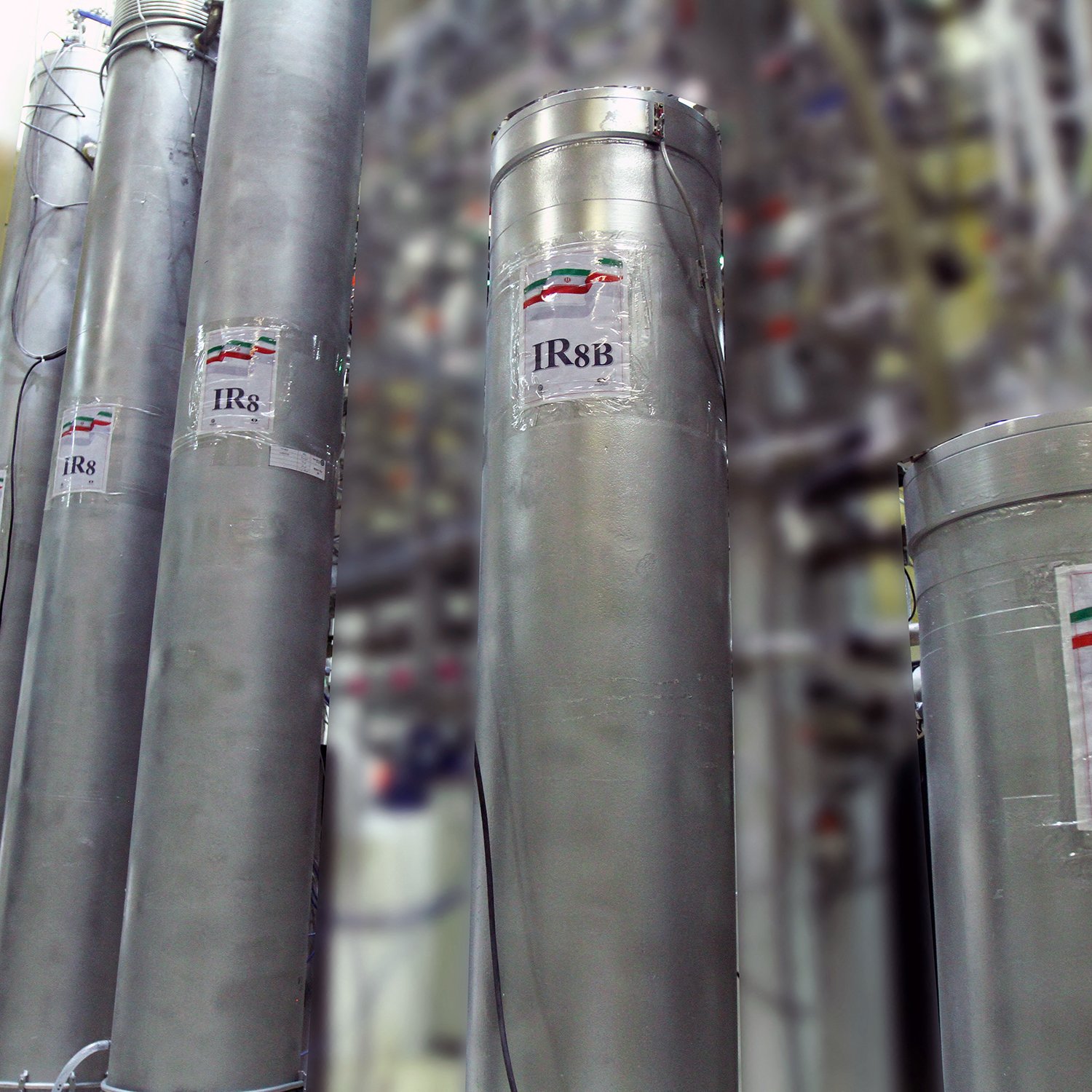 تظهر في الصورة مجموعة من الأسطوانات المعدنية، وكل منها يحمل ملصق مكتوب عليه "IR8" أو "IR8s" مع رمز علم دولة إيران. خلف هذه الأسطوانات، يمكن رؤية مجموعة من الأنابيب والتجهيزات الأخرى، مما يشير إلى أنها قد تكون جزءًا من منشأة صناعية أو مركز بحث علمي. الجو العام في الصورة يعكس بيئة تقنية عالية ومجهزة بأجهزة متطورة.