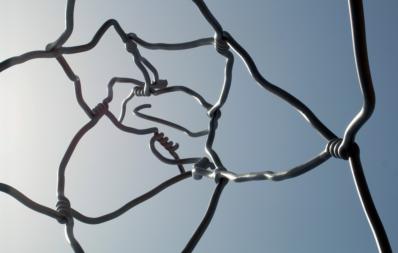 L'image montre une structure artistique composée de fils métalliques entrelacés. Les fils sont courbés et forment des nœuds à certains endroits, créant un motif complexe et abstrait. On peut voir le ciel en arrière-plan, ce qui met en valeur les formes des fils. L'ensemble dégage une impression de légèreté et de fluidité, tout en étant à la fois structurée et organique.