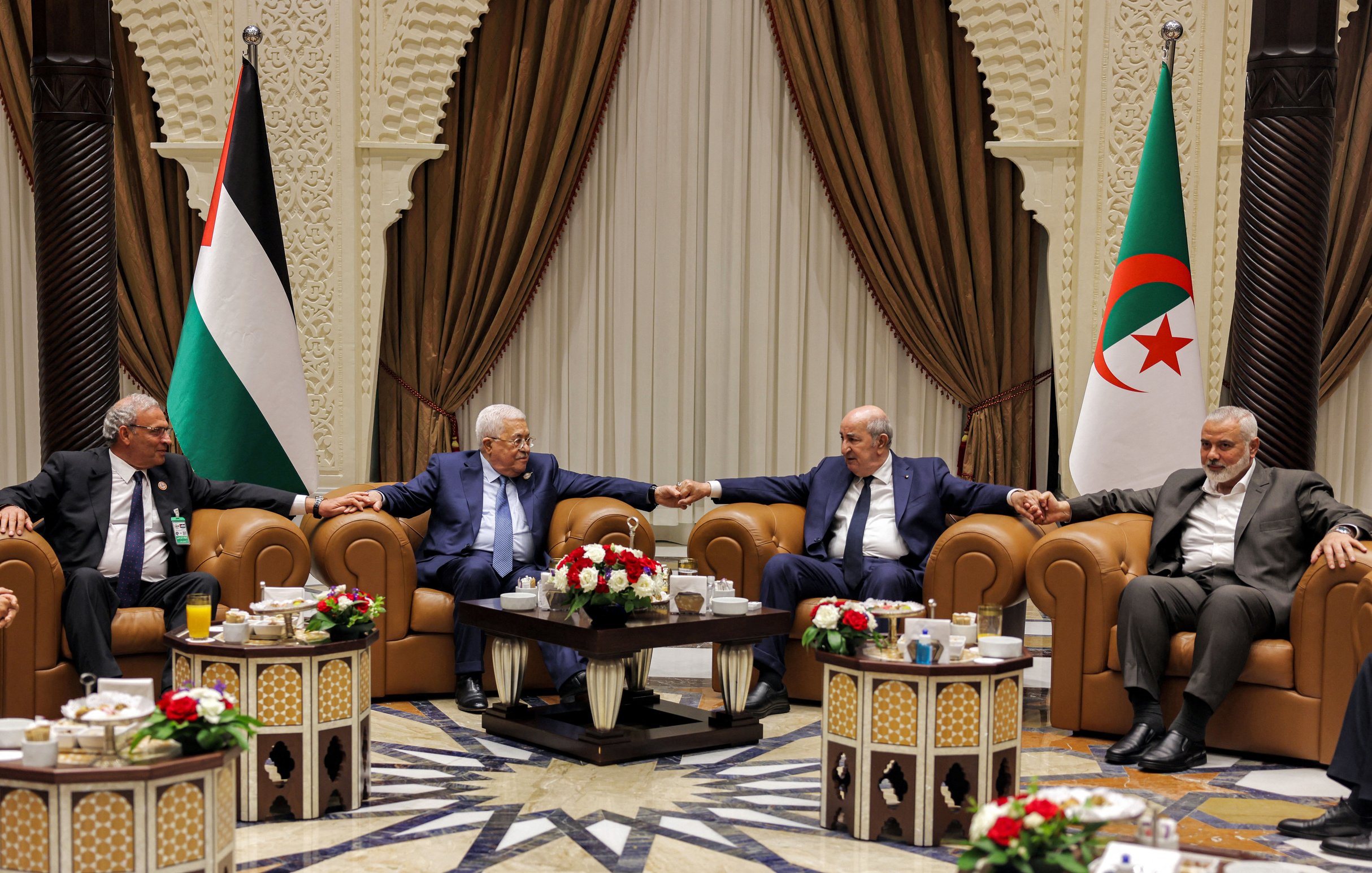 تظهر الصورة مجموعة من الأشخاص seated في غرفة مزينة بشكل أنيق، حيث يجلس أربعة رجال على أرائك مريحة. بينهم أجواء من التعاون والود، إذ يتبادلون الحديث مع قيامهم بتشابك الأيادي. في الخلفية، توجد أعلام فلسطين والجزائر، ما يشير إلى وجود موضوع يتعلق بالتعاون العربي أو القضايا السياسية بين البلدين. الطاولات أمامهم مزينة بتنسيقات من الزهور ومشروبات. الأجواء تبدو رسمية ولكن ودية في الوقت نفسه.