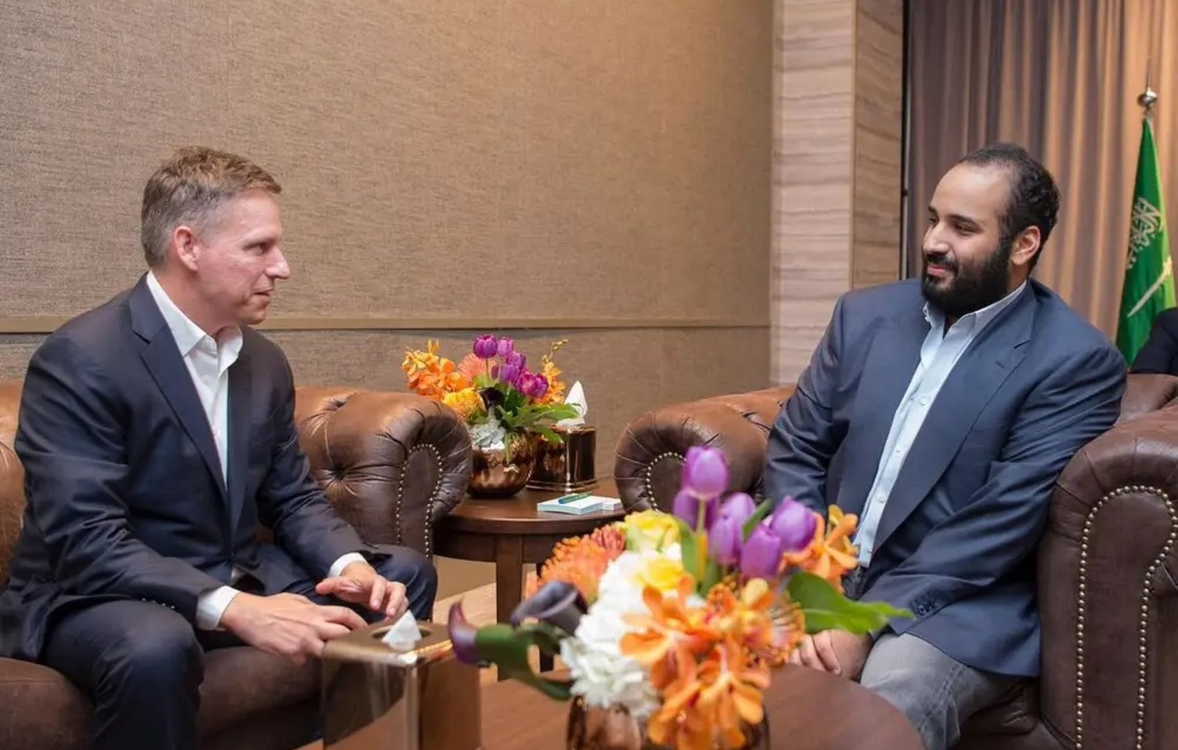 Two men in suits sit in a room, conversing near a floral arrangement on a table.