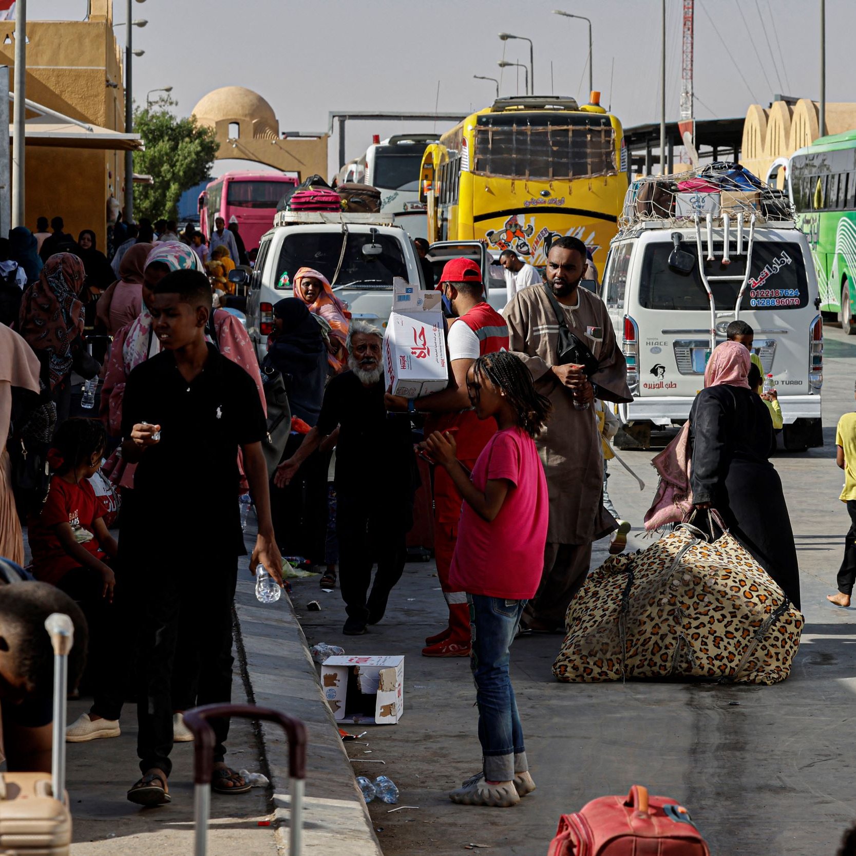 در تصویر، یک ایستگاه اتوبوس یا ترمینال مشاهده می‌شود که در آن تعدادی اتوبوس در حال توقف هستند. مردم زیادی در حال رفت و آمد هستند، برخی در حال حمل چمدان و بارهای خود هستند. در میان این افراد، کودکان نیز دیده می‌شوند که در حال بازی یا حرکت هستند. برخی از افراد در حال استراحت و نشستن بر روی ساک‌های خود هستند. فضای ترمینال به نظر شلوغ و پرجنب و جوش می‌آید و آفتاب روشن در حال تابش است.