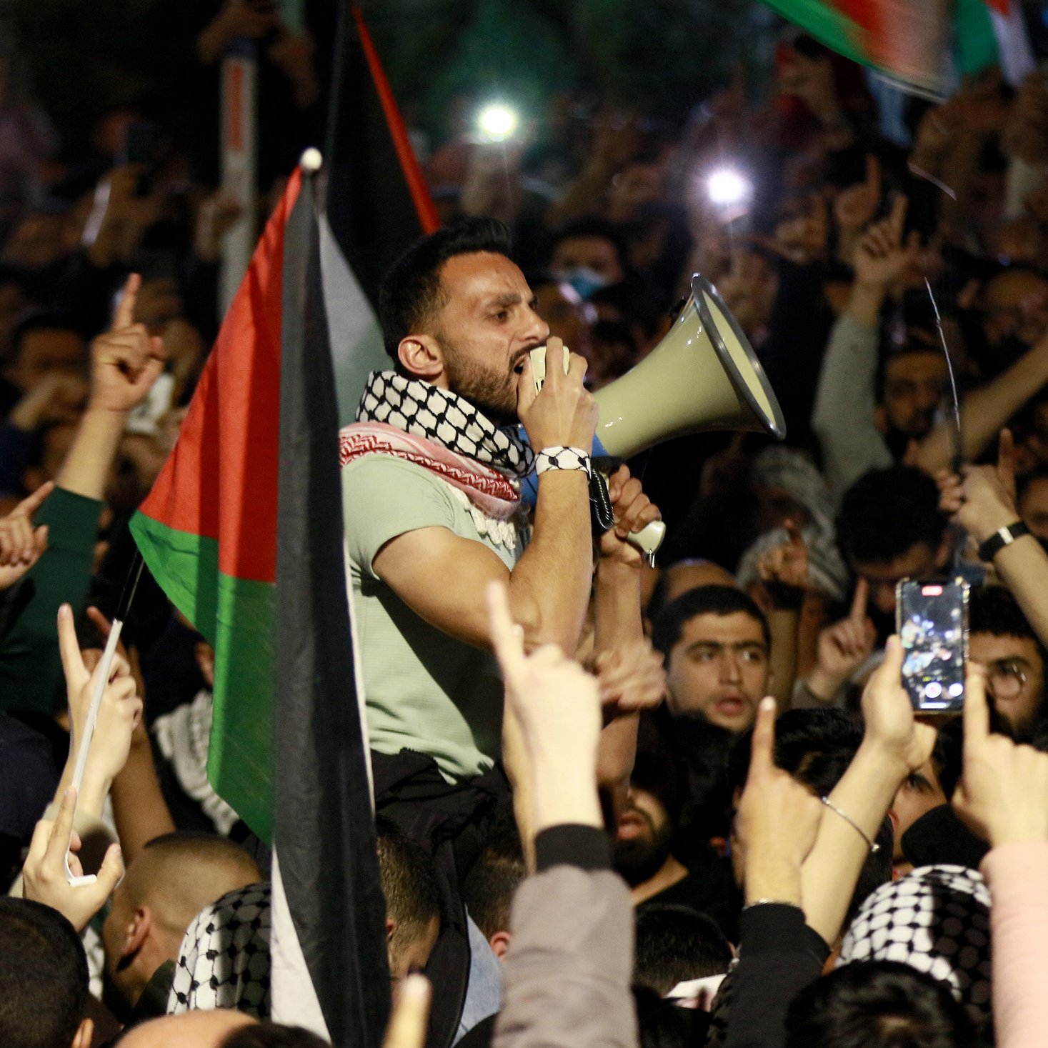 L'image montre un grand rassemblement de personnes, probablement lors d'une manifestation. Au premier plan, un homme utilisant un mégaphone pour parler à la foule. Il est entouré de nombreux participants qui lèvent les bras et brandissent des drapeaux, notamment des drapeaux palestiniens. L'atmosphère semble électrisante et engagée, avec des gens qui filment l'événement avec leurs téléphones. La scène dénote une forte mobilisation et un sentiment collectif.
