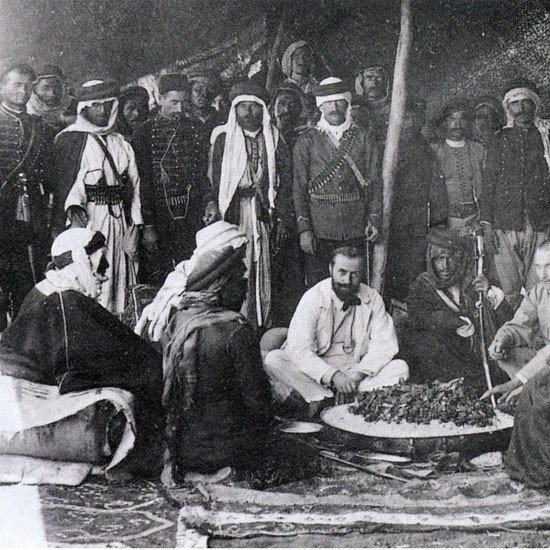 این تصویر تاریخی گروهی از مردان را در یک محیط سنتی نشان می‌دهد. آنها در یک چادر یا فضای باز گرد هم آمده‌اند و به نظر می‌رسد در حال تهیه یا صرف غذا هستند. چند نفر از آن‌ها لباس‌های سنتی عربی به تن دارند، از جمله عبا و headscarf (کشیه). در مرکز تصویر، چند نفر در حال نشستن و گفتگو هستند و سایرین ایستاده یا نشسته در اطراف آن‌ها قرار دارند. فضای تصویر گرم و صمیمی به نظر می‌رسد و نمایانگر فرهنگی غنی و سنت‌های اجتماعی است.