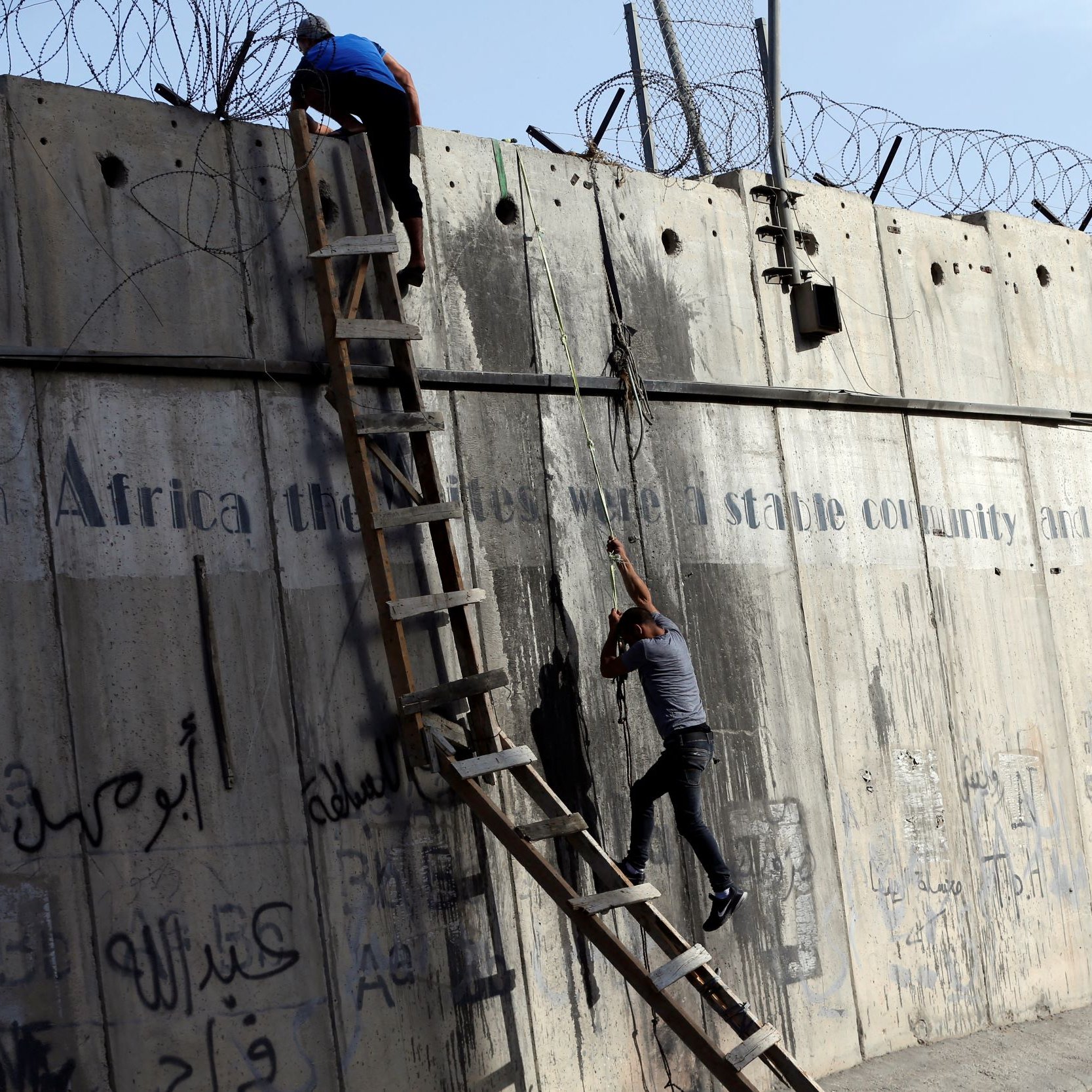 L'immagine mostra due persone che stanno scalando un muro di cemento, sul quale è presente della rete metallica. Dietro di loro, è visibile una scala che qualcuno sta utilizzando per salire. Il muro è coperto da graffiti e scritte, in varie lingue, che sembrano affrontare temi legati alla storia e alla condizione sociale. La scena evoca un senso di tensione e disperazione, con le persone che tentano di superare un ostacolo fisico, simbolo di divisione.
