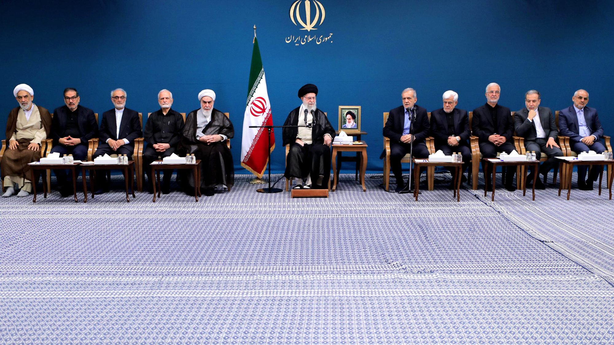 الصورة تظهر مجموعة من الأشخاص يجلسون في صفين. في الخلفية يوجد علم إيران، وهناك صورة للمرشد الأعلى. الأفراد يرتدون ملابس رسمية، ويبدو أنهم في مناسبة أو اجتماع رسمي. الغرفة تتميز بجو رسمي ومهيب، مع سجادة تغطي الأرض.