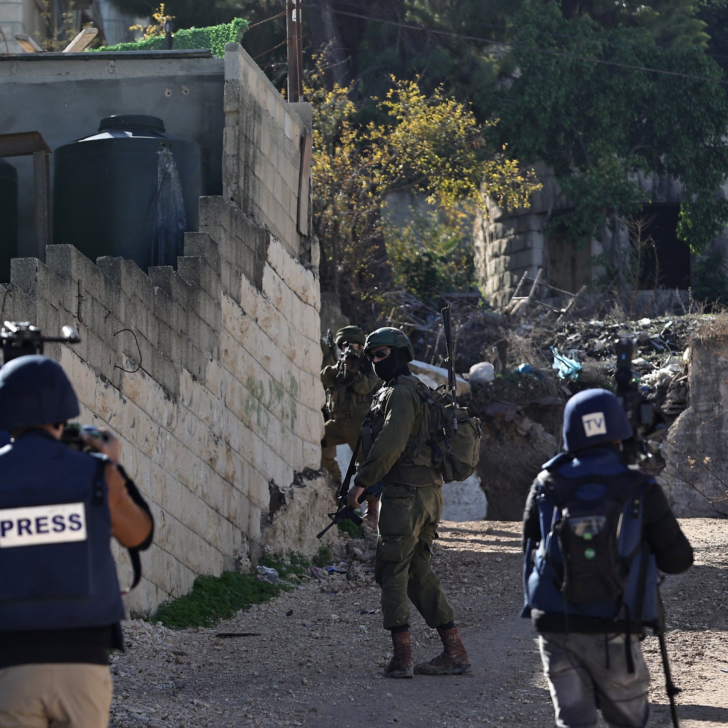 The image depicts a scene in an urban environment, likely involving a military or conflict situation. In the foreground, there are three individuals wearing vests labeled "PRESS," indicating they are journalists or reporters. They appear to be observing or documenting the situation. In the background, a soldier in military gear is positioned, holding a weapon and looking in the direction of the journalists. The surroundings include a wall, some buildings, and debris, suggesting the area may be experiencing instability or conflict.