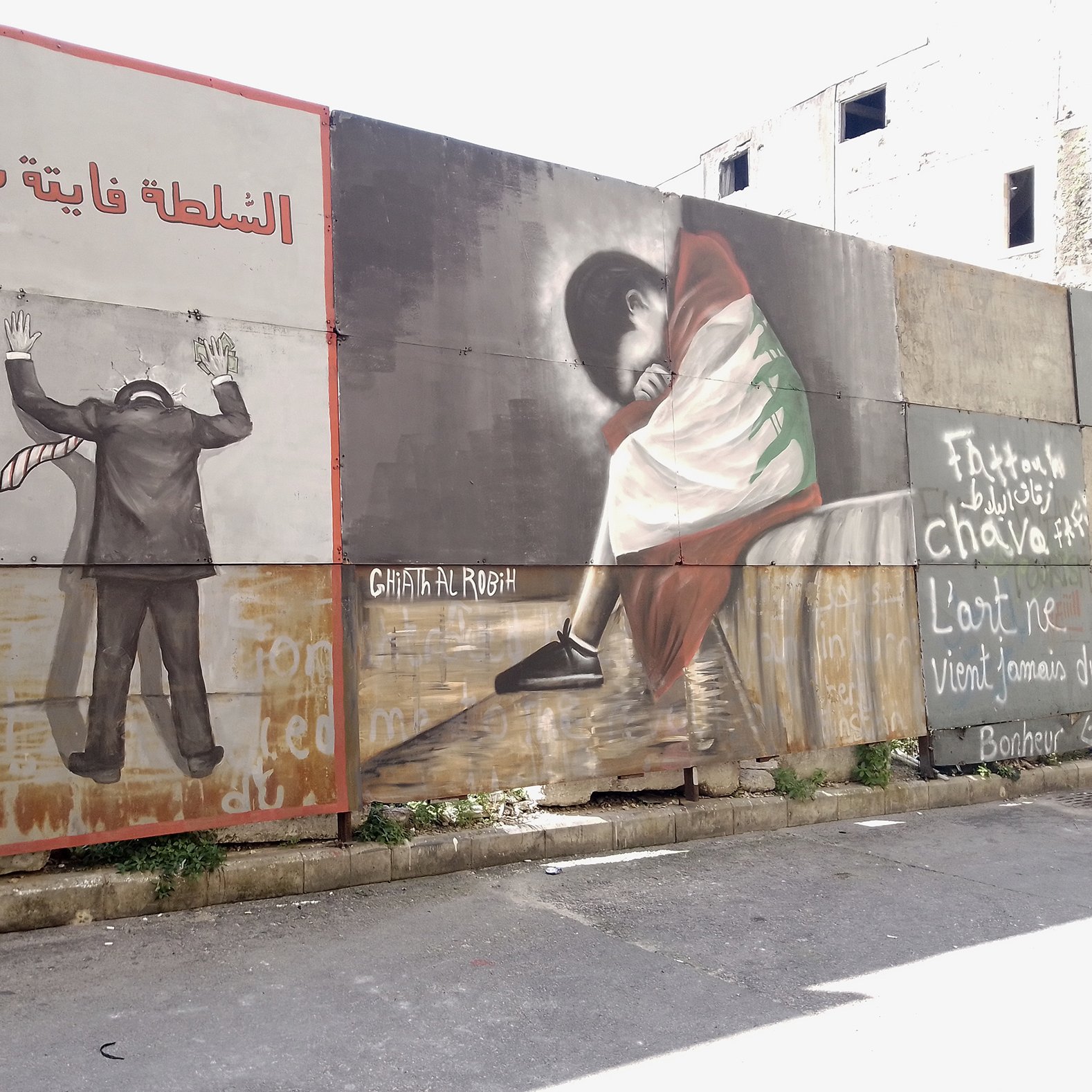 L'image montre un mur recouvert de plusieurs peintures murales. À gauche, on voit une figure humaine en costume, les mains levées, comme en signe de désespoir ou de soumission, avec une inscription en arabe qui pourrait évoquer des thèmes de répression. À droite, une autre figure, probablement d'une personne en train de pleurer ou de s'asseoir tristement, est représentée, vêtue d'une robe aux couleurs d'un drapeau, ce qui pourrait symboliser une forte connexion à une identité culturelle ou nationale. Le fond comporte également des mots ou des phrases en français et en arabe, ajoutant une dimension de commentaire social ou politique à l'œuvre. L'ensemble évoque des thèmes de lutte, d'identité et d'émotion.