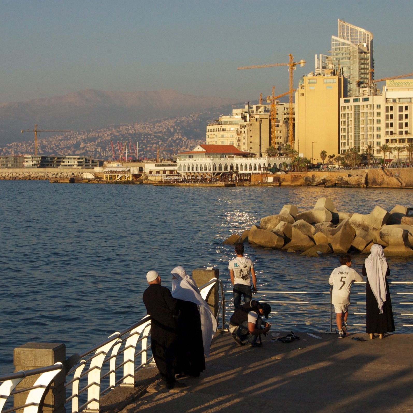L'image montre un paysage côtier au bord de la mer. On peut voir une promenade avec des personnes qui se tiennent près de l'eau, probablement en train de profiter du coucher de soleil. Au fond, il y a des bâtiments modernes et quelques grues de construction, suggérant un développement urbain. La mer est calme et reflète la lumière dorée du soleil. La scène dégage une atmosphère paisible et agréable.