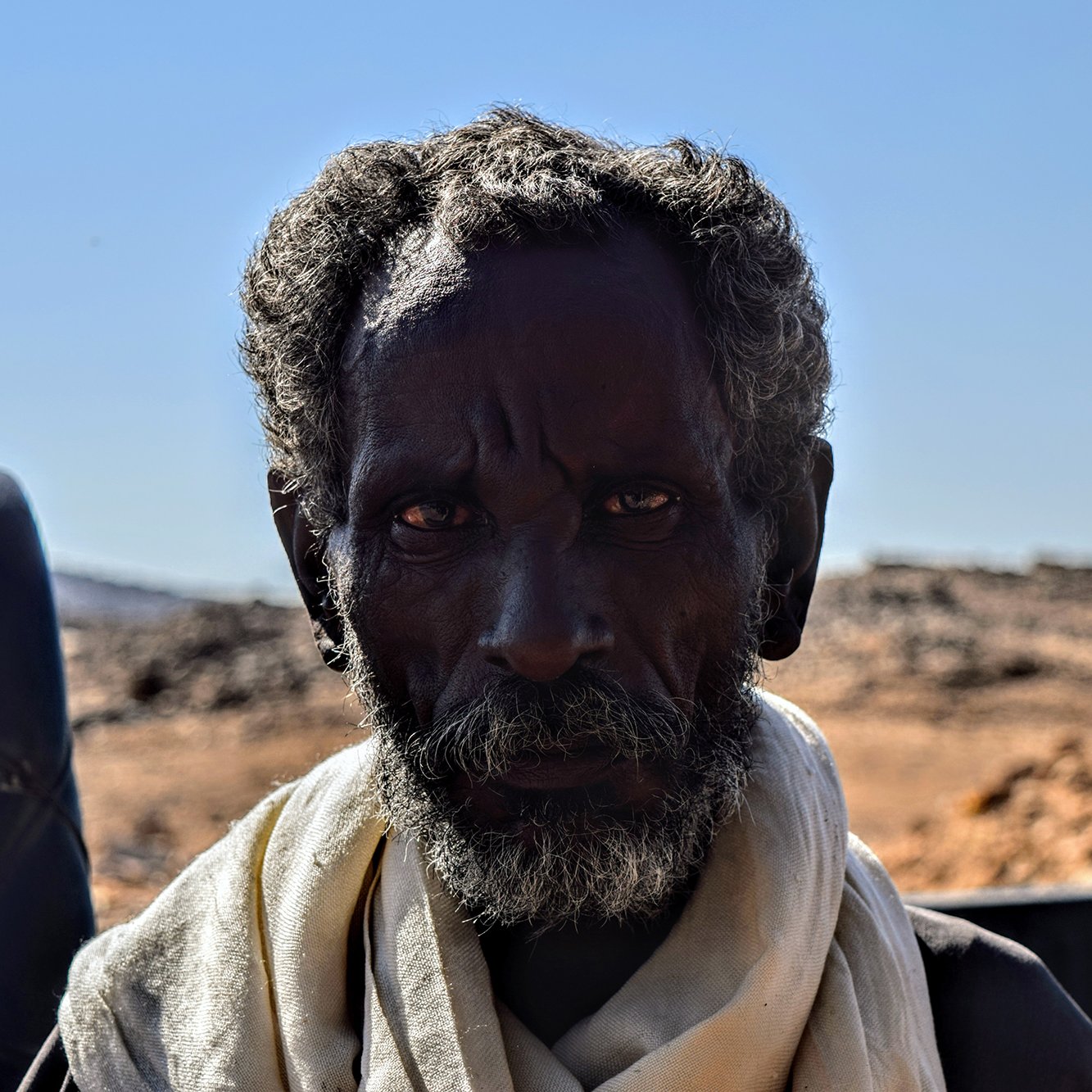 The image features an elderly man with dark skin and a thick, gray beard. He has deep-set eyes that convey a sense of wisdom and experience. The background is blurred but suggests a rugged landscape, likely arid or rocky, with a clear blue sky. The man is wearing a light-colored scarf around his neck. His expression appears serious and contemplative.