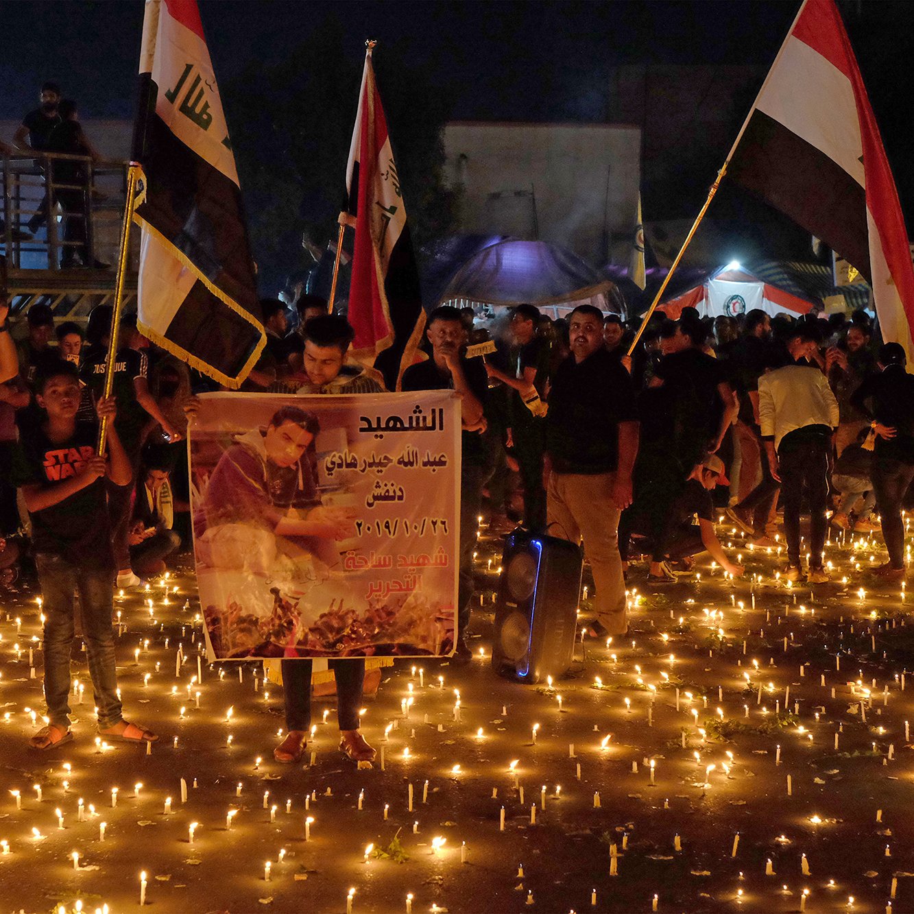 L'image montre une foule rassemblée dans une rue sombre, illuminée par des centaines de bougies. Les personnes portent des affiches et des banderoles, certaines mettant en avant des portraits ou des messages en hommage à des individus, probablement des victimes ou des figures importantes. Il y a aussi des drapeaux en arrière-plan, suggérant un contexte national. L'atmosphère semble être à la commémoration ou au rassemblement pour une cause, avec un sentiment de solidarité visible parmi les participants.
