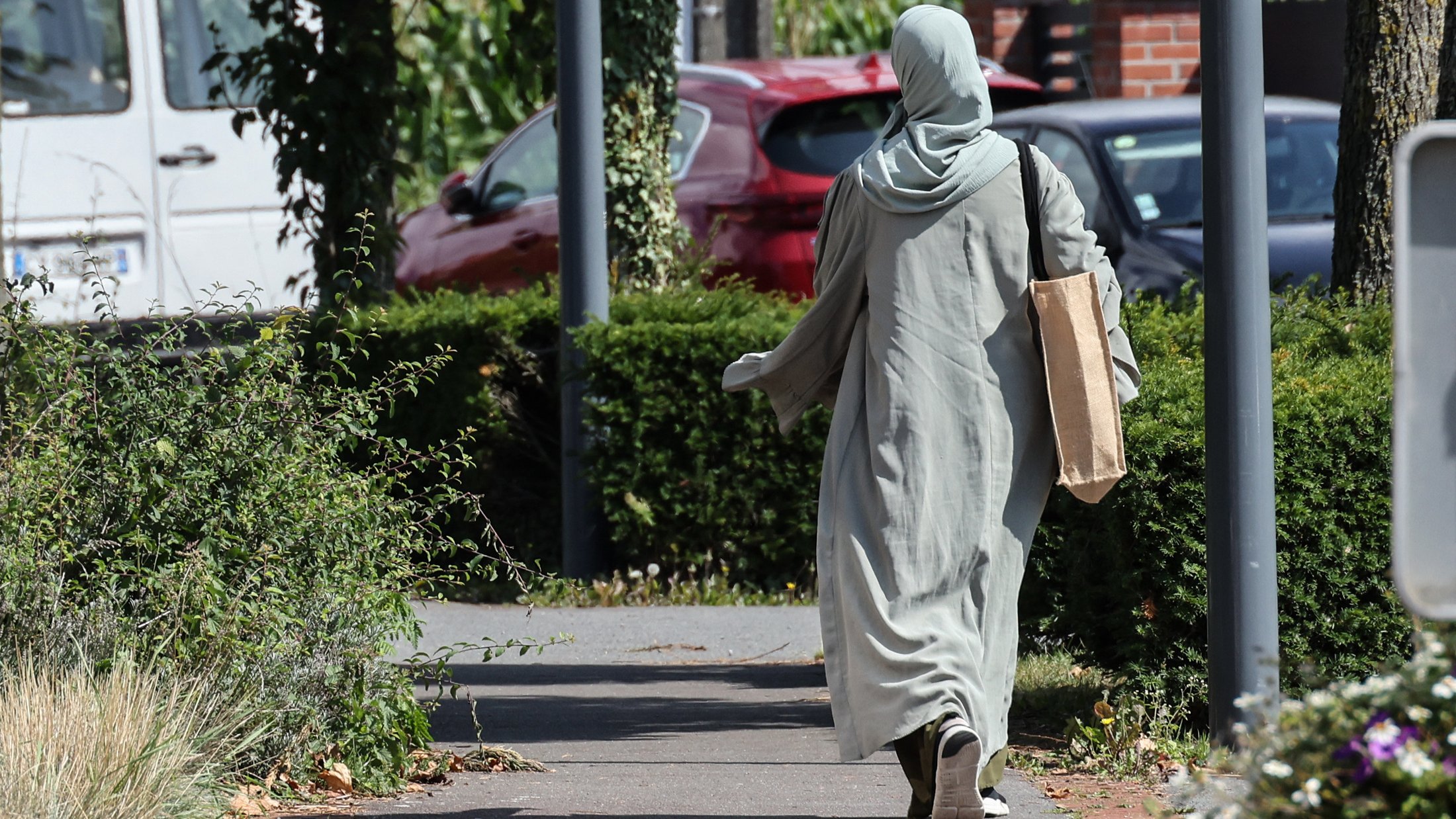 L'image montre une personne marchant sur un chemin. Elle porte une longue robe de couleur claire et un foulard. Elle tient un grand sac en papier dans une main. Le décor environnant inclut des buissons et des arbres, avec des voitures en arrière-plan. La scène semble tranquille et ensoleillée.