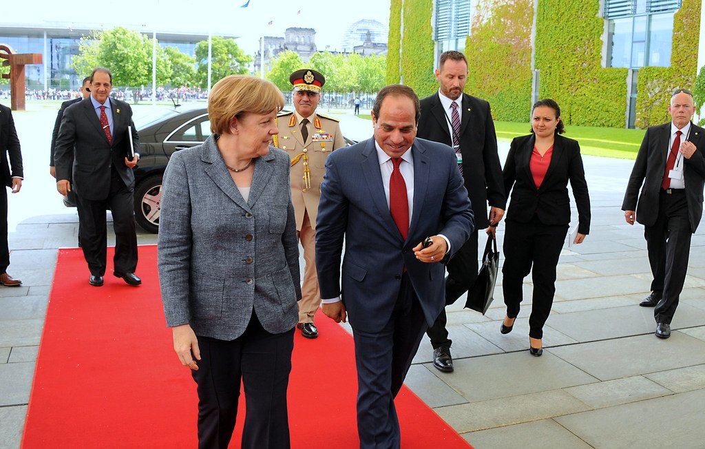 L'image montre un moment officiel, probablement lors d'une rencontre diplomatique. Une femme, portant un blazer gris, marche sur un tapis rouge aux côtés d'un homme en costume sombre. Ils semblent engagés dans une conversation. À l'arrière-plan, on peut voir des personnes en costume, ainsi que des bâtiments modernes et une végétation. L'atmosphère est formelle et professionnelle.