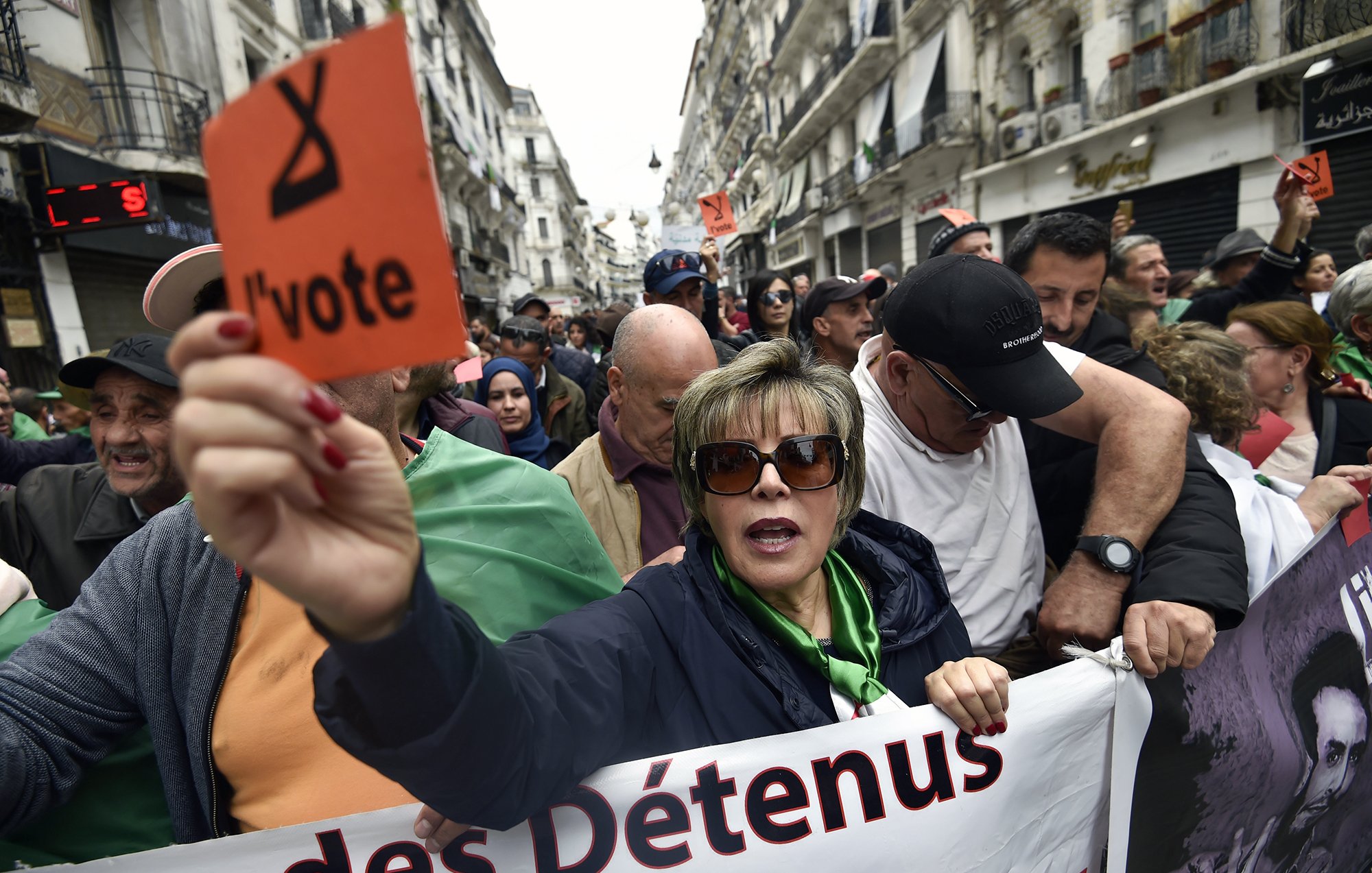 The image depicts a crowd of protesters gathered in an urban setting, likely during a demonstration. A woman in the foreground is holding an orange sign that reads "l'vote" while wearing sunglasses and a green top. The crowd behind her appears to be a mix of men and women, some holding signs and banners. The atmosphere conveys a sense of urgency and solidarity among the protesters, who seem to be advocating for a political cause. The background includes urban architecture, suggesting a city environment.