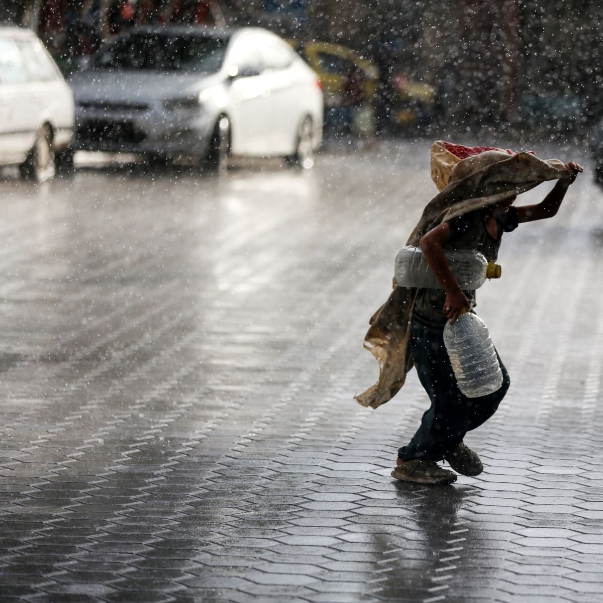 L'image montre un enfant courant sous la pluie intense, portant une écuelle en métal et un sac. Il semble se protéger de la pluie avec un tissu qui couvre sa tête et ses épaules. Le sol est mouillé et réfléchit la lumière, tandis que l'environnement autour de lui est flou en raison des cordes de pluie. Des voitures sont visibles en arrière-plan, accentuant l'atmosphère urbaine de la scène.