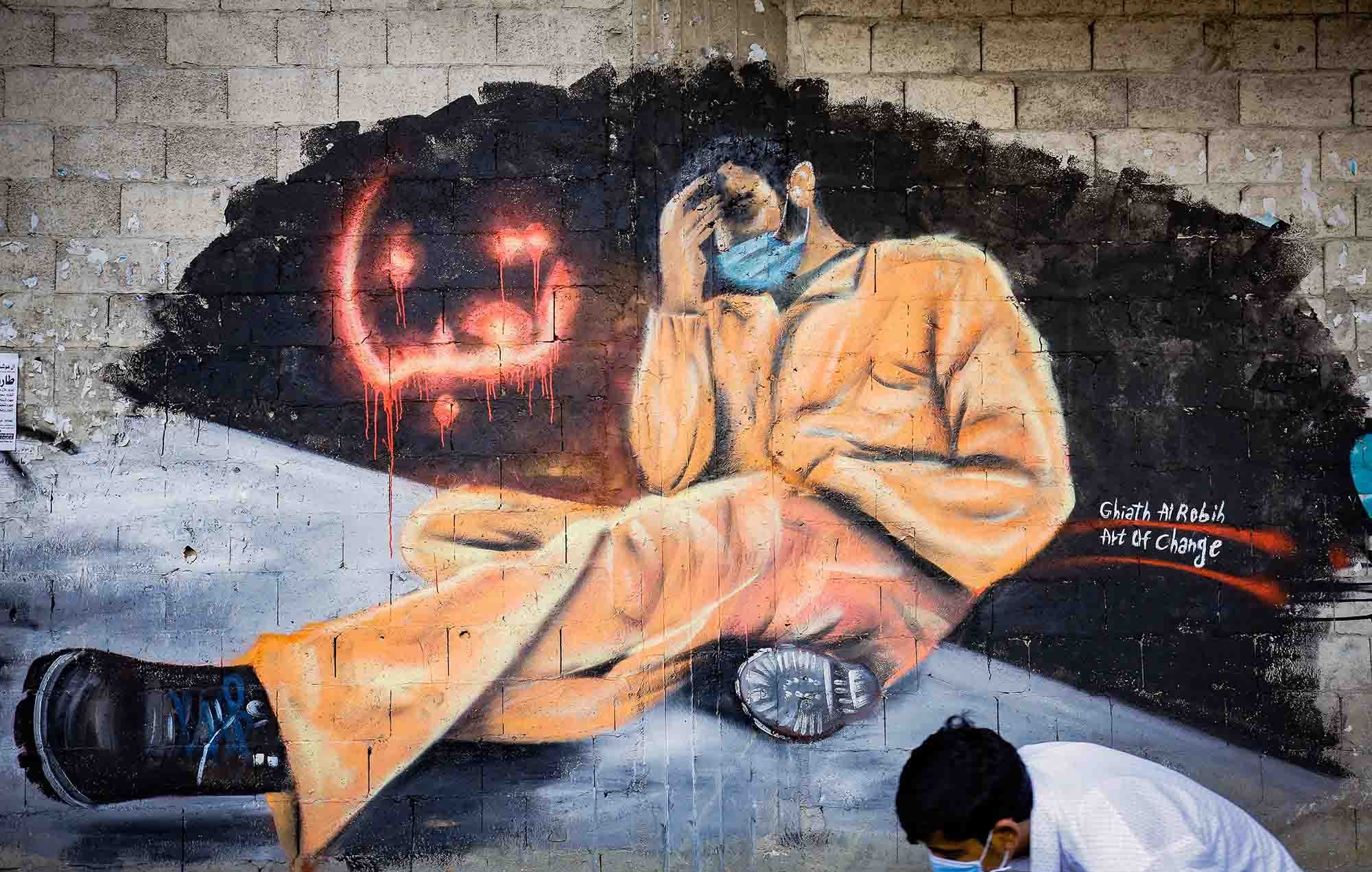 L'immagine mostra un murales che rappresenta una figura umana seduta, vestita con una tuta arancione, che tiene la testa tra le mani in un gesto di disperazione o riflessione. Il personaggio sembra essere in una posizione di vulnerabilità e solitudine. Accanto a lui, ci sono scritte in arabo e gocce di colore rosso che aggiungono un senso di drammaticità. Nella parte inferiore della scena, si vede un ragazzo in posizione inginocchiata, che sembra osservare il murales. Questo opera sembra voler trasmettere un messaggio profondo sulla sofferenza e l'oppressione.
