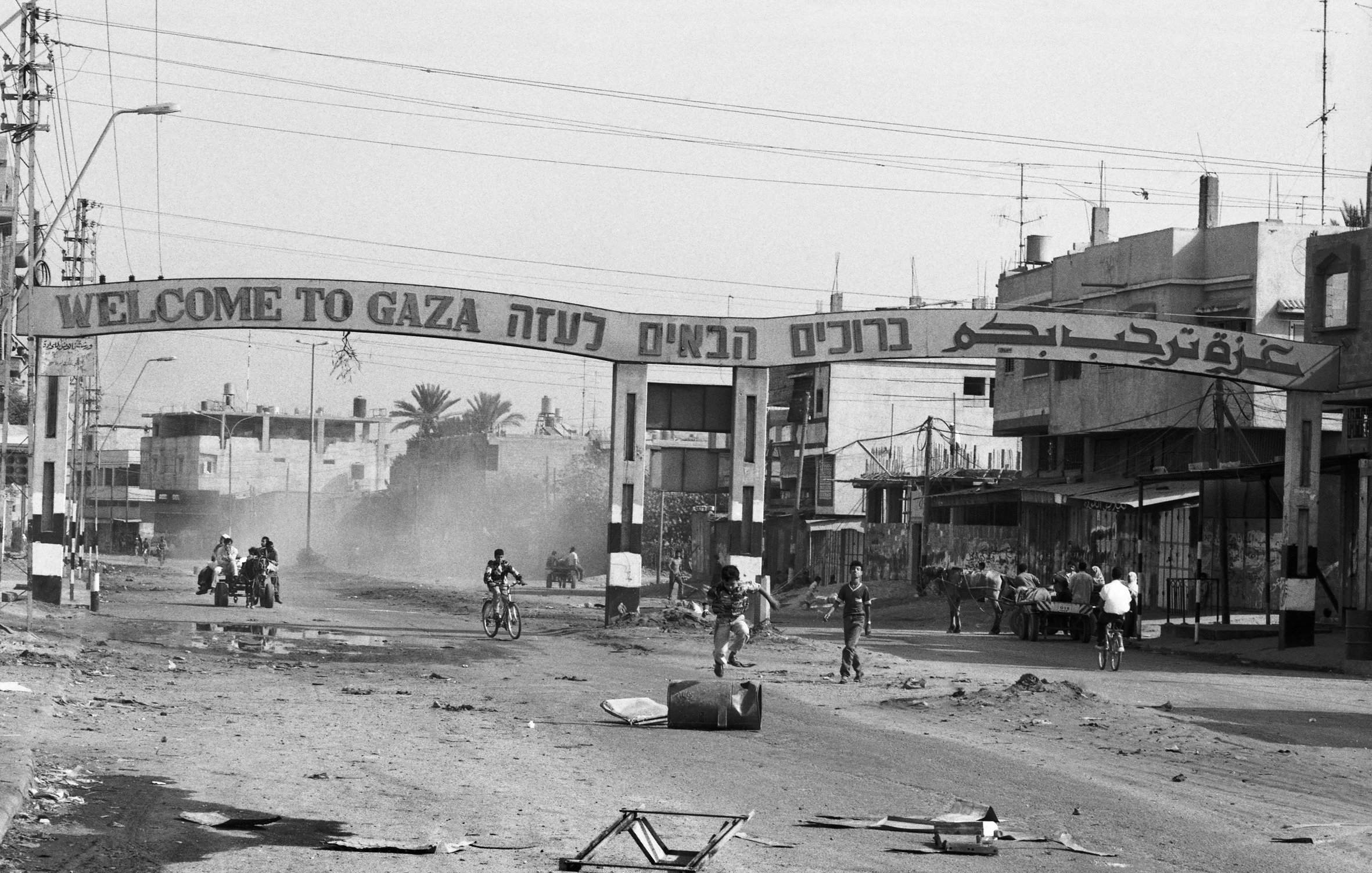 L'image montre une rue dans une ville, probablement à Gaza, avec une grande arche à l'entrée où il est écrit "Welcome to Gaza" en plusieurs langues. La scène est en noir et blanc, ce qui donne une atmosphère nostalgique ou dramatique. On aperçoit quelques personnes sur des bicyclettes et d'autres regroupées sur le côté de la route. Le décor urbain semble en désordre, avec des bâtiments qui montrent des signes d'usure et de dévastation. De la poussière s'élève, ajoutant à l'ambiance de la scène.