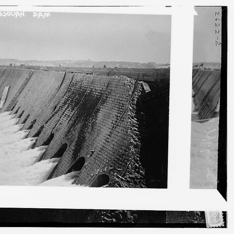 L'image représente un barrage, probablement le barrage d'Assouan, avec une structure en béton. On peut voir de l'eau s'écoulant à travers des ouvertures sur le côté du barrage. Le paysage environnant semble naturel, et le ciel est clair. L'image évoque une impression de puissance hydraulique et d'ingénierie.