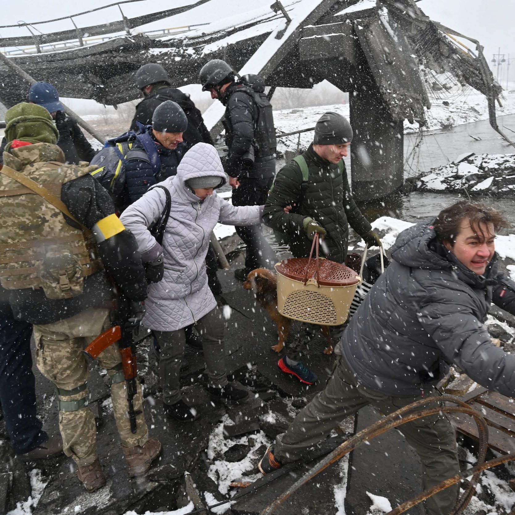 تظهر الصورة مجموعة من الأشخاص في منطقة مدمرة، حيث يبدو أن هناك أضرار جسيمة في البيئة المحيطة. يُشاهد بعض الأفراد يرتدون ملابس عسكرية، بينما الآخرين يرتدون ملابس عادية ويمسكون بأغراض مختلفة. الثلوج تتساقط بكثافة، مما يُعطي انطباعًا عن ظروف مناخية قاسية. يبدو أن الأشخاص يبذلون جهدًا في المساعدة أو التحرك بأمان في هذه البيئة، مما يشير إلى حالة طوارئ أو أزمة.