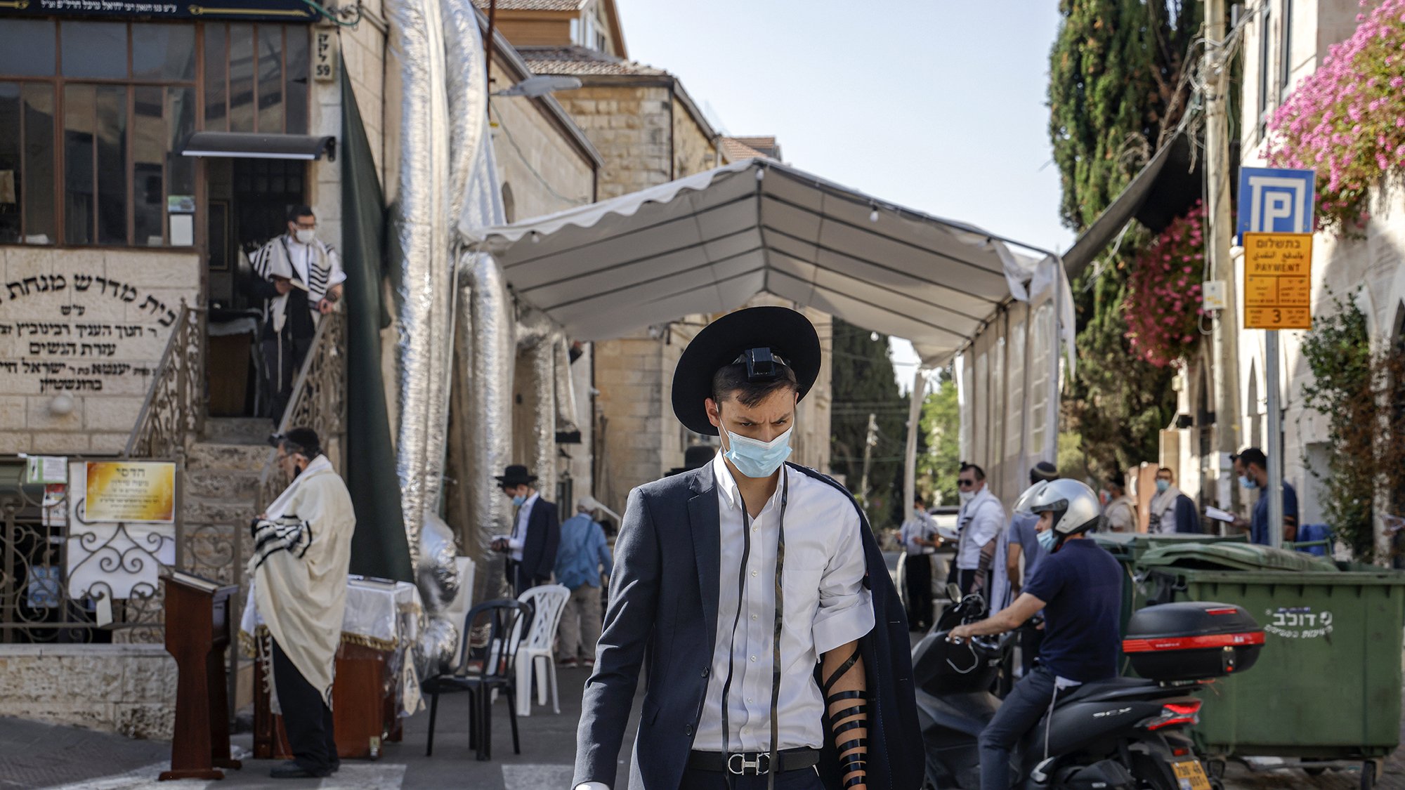 تظهر الصورة شابًا يرتدي بدلة مع قبعة، وهو يحمل هاتفًا محمولًا في يده. يتزين ذراعه بلفائف التوراة (تيفيلين)، ويبدو أنه في أحد الأماكن العامة. في الخلفية، توجد مجموعة من الأشخاص، وبعضهم يرتدي ملابس تقليدية، كما يظهر خيمة كبيرة على جانب الطريق. تتميز الصورة بأجواء حضرية مع وجود نباتات وبيئات سكنية.