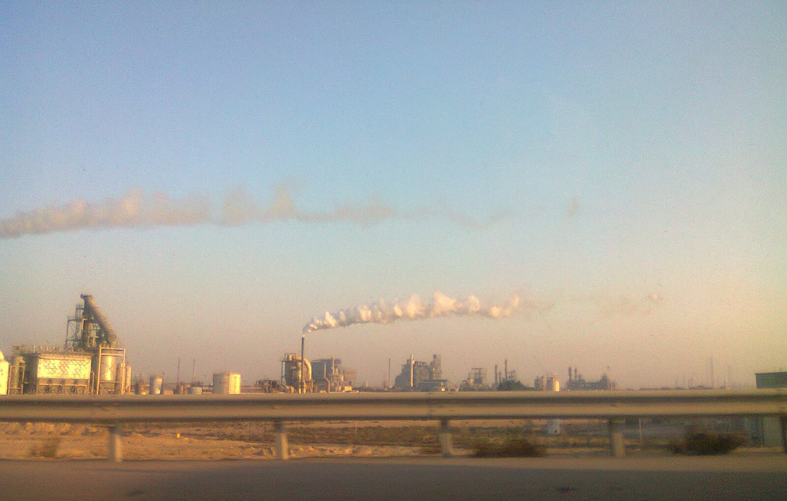 L'image montre un paysage industriel avec plusieurs structures d'usine. On voit de grandes cheminées qui émettent des nuages de fumée blanche dans le ciel. Le ciel est clair, avec une lumière douce, ce qui donne une atmosphère tranquille malgré l'activité industrielles. Au premier plan, on aperçoit une route avec une barrière en béton, indiquant que l'on observe ce paysage en passant en véhicule. L'ensemble dégage une impression de production industrielle dans un environnement ouvert.