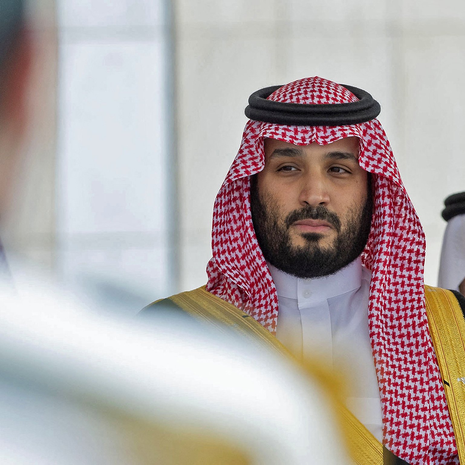 L'image montre un homme portant des vêtements traditionnels saoudiens, y compris un keffieh et un thobe. Il a une expression sérieuse et regarde vers la caméra ou un point de vue adjacent. Son entourage semble formel, avec une ambiance de cérémonie ou de rencontre officielle. Les couleurs dominantes sont les tons de rouge et de blanc du keffieh, ainsi que des touches dorées sur sa tenue.
