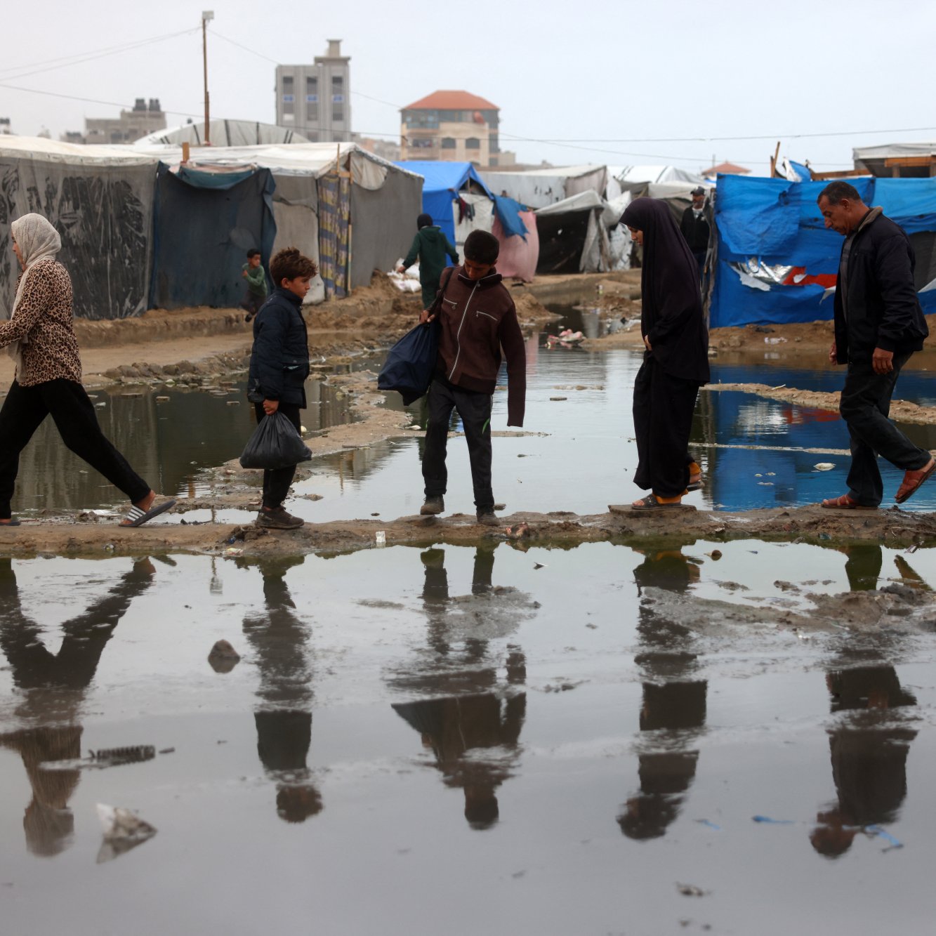 L'image montre des personnes marchant à travers un camp de tentes, avec des surfaces détrempées et des flaques d'eau. On peut voir une femme en hijab, un enfant, et plusieurs adultes, tous avançant sur des passages surélevés ou des morceaux de terrain au-dessus de l'eau. L'environnement semble difficile, et les refuges sont constitués de bâches et de tissus. Les reflets des marcheurs se voient dans les flaques, ajoutant une dimension visuelle à la scène.