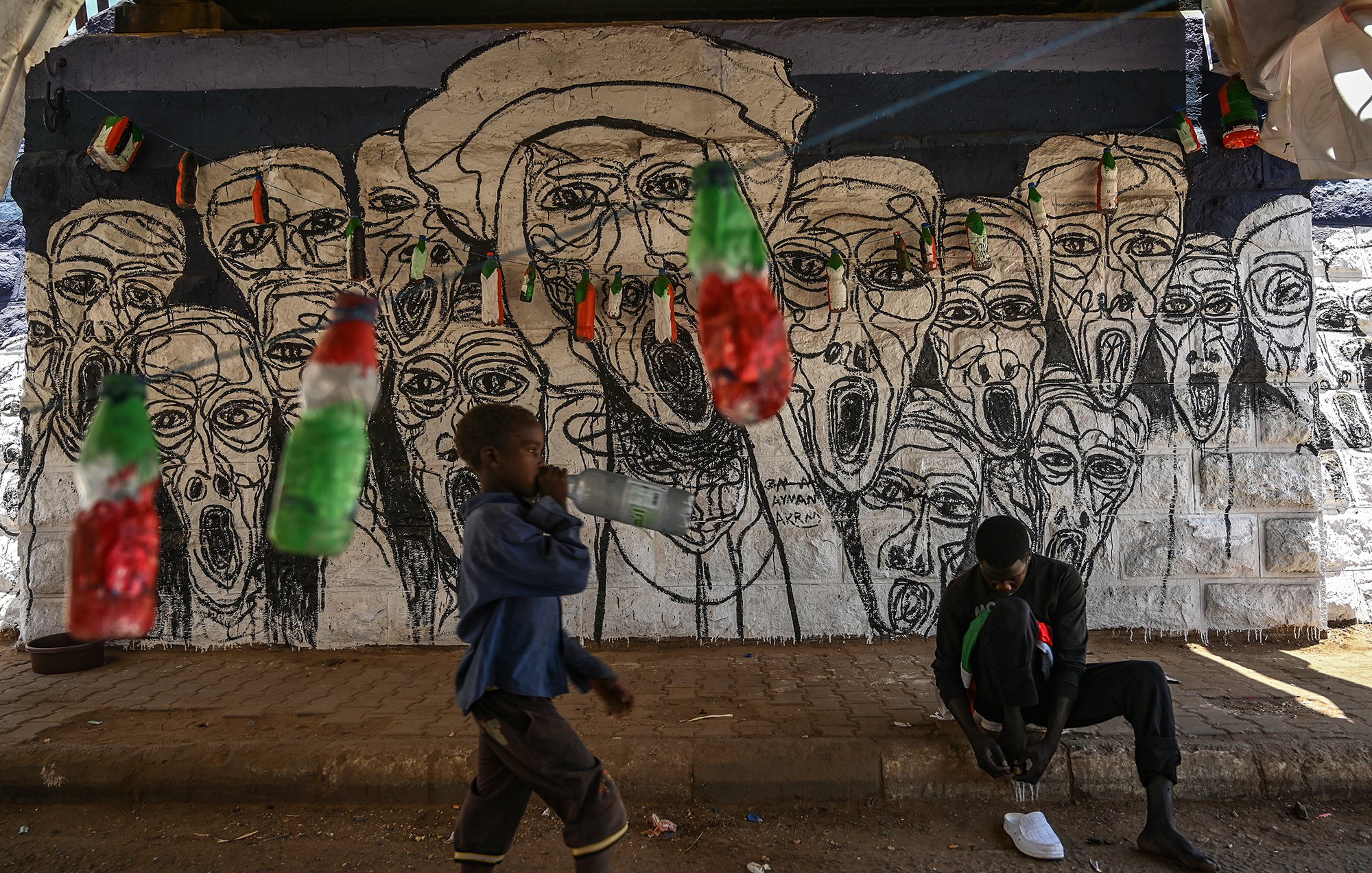 L'image montre un mur peint en arrière-plan, où se trouvent des visages aux traits exagérés, tous avec des expressions de cri, créant une atmosphère vibrante et expressive. Devant ce mur, un enfant marche en tenant une bouteille, tandis qu'un autre individu est assis au sol, occupé à lacer ses chaussures. Des bouteilles colorées, probablement en plastique, sont suspendues au-dessus d'eux, ajoutant une touche de couleur à la scène. Cette image reflète un environnement urbain dynamique, mêlant art de rue et vie quotidienne.