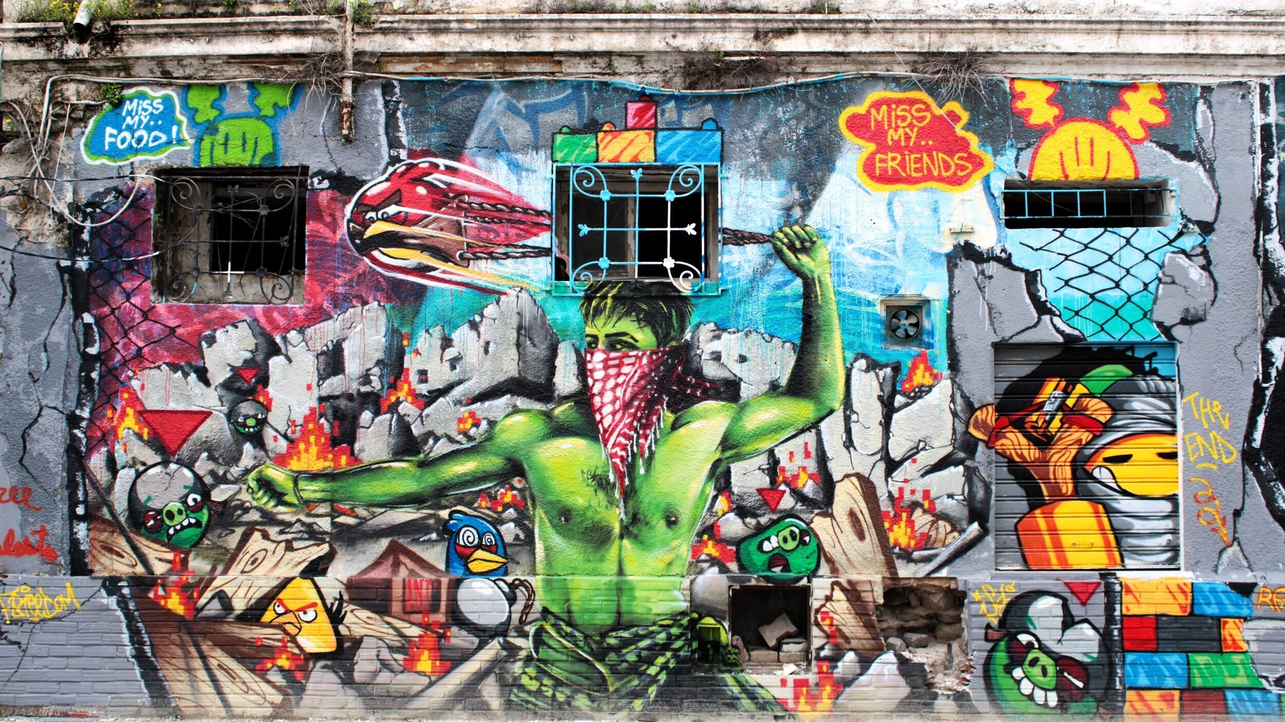 L'immagine mostra un murales colorato su un muro esterno. Al centro, c'è un personaggio verde con un fazzoletto rosso, che sembra esprimere un forte senso di protesta o di resistenza. Sullo sfondo, si possono notare elementi di graffiti e illustrazioni vivaci, tra cui simboli e oggetti in movimento, che creano un'atmosfera dinamica e urbana. Il murales combina diverse tecniche artistiche, rendendolo molto interessante visivamente.