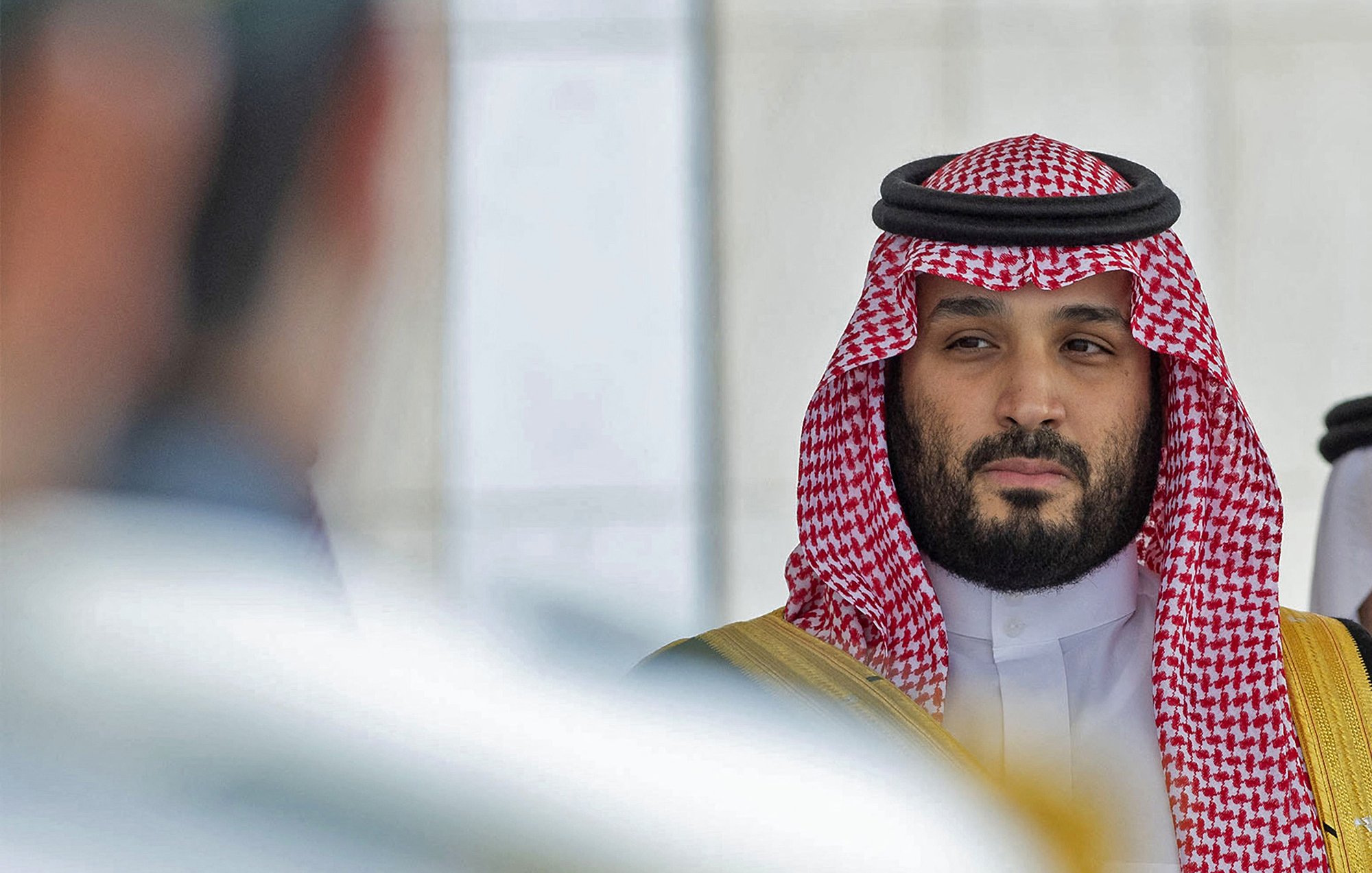 L'image montre un homme portant des vêtements traditionnels saoudiens, y compris un keffieh et un thobe. Il a une expression sérieuse et regarde vers la caméra ou un point de vue adjacent. Son entourage semble formel, avec une ambiance de cérémonie ou de rencontre officielle. Les couleurs dominantes sont les tons de rouge et de blanc du keffieh, ainsi que des touches dorées sur sa tenue.
