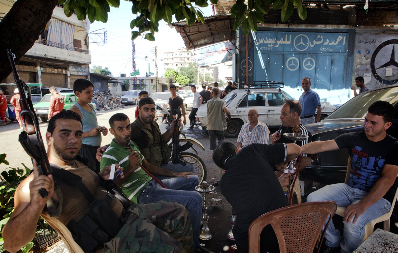 L'image montre un groupe d'hommes assis dans une rue animée. Certains d'entre eux portent des vêtements décontractés et semblent discuter ou se détendre ensemble. On peut voir un homme armé avec une arme à feu, ce qui suggère une ambiance tendue ou de conflit. En arrière-plan, il y a des voitures et des bâtiments, ainsi qu'une enseigne avec le logo d'une marque automobile. L'atmosphère semble informelle, malgré la présence d'armes.
