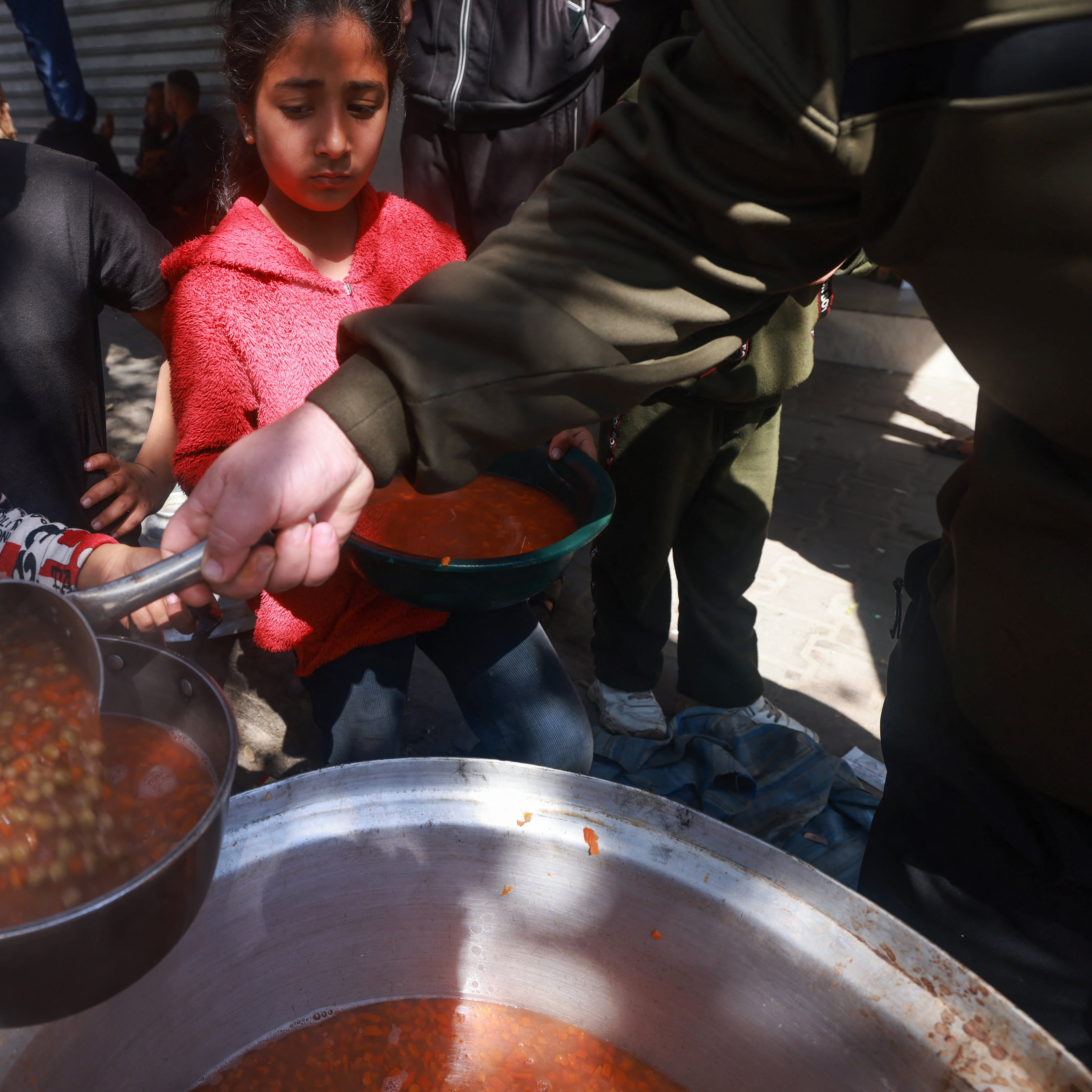 در این تصویر، گروهی از کودکان در حال دریافت غذا هستند. یکی از کودکان که لباس‌های رنگارنگ دارد، با دقت در حال ریختن خورش یا سوپ به ظرفش است. در پس‌زمینه، سایر کودکان نیز منتظر هستند تا نوبتشان برسد. محیط بیرونی به نظر شلوغ و غیررسمی است و نشان‌دهنده یک فعالیت جمعی برای سرو غذا به افراد نیازمند می‌باشد.