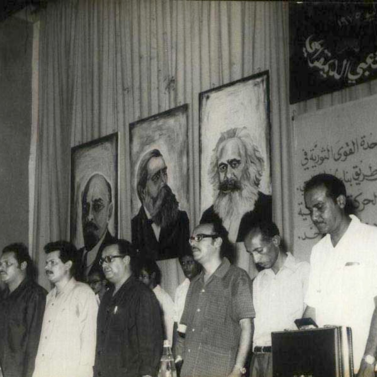 L'image montre un groupe d'hommes se tenant debout en ligne, probablement lors d'un événement ou d'une conférence. Ils semblent pris dans un moment de réflexion ou de recueillement. À l'arrière-plan, on peut voir plusieurs portraits accrochés au mur, représentant des figures historiques ou intellectuelles, dont certaines sont bien connues. Le décor suggère un contexte politique ou idéologique, avec des inscriptions en arabe sur le mur qui renforcent ce thème. L'atmosphère générale semble sérieuse et engagée.