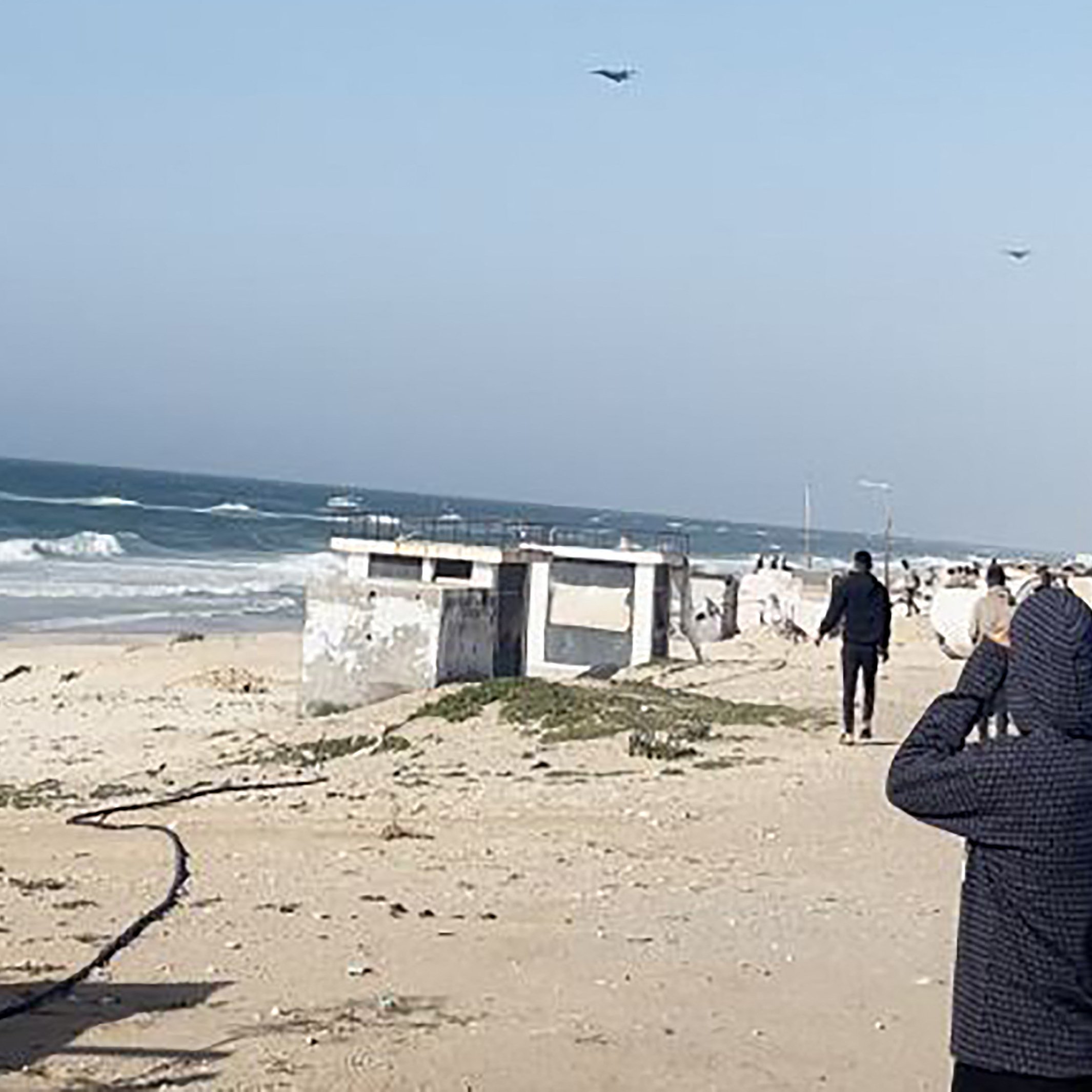 L'image montre une scène côtière avec des vagues de mer s'écrasant sur le rivage. Au premier plan, plusieurs personnes regardent vers l'horizon, peut-être fascinées par quelque chose dans le ciel. On peut également voir des véhicules stationnés sur le côté. L'ambiance semble calme, avec un ciel dégagé et une légère brise. La plage est principalement sablonneuse, et quelques éléments tels que des algues sont visibles sur le sol.