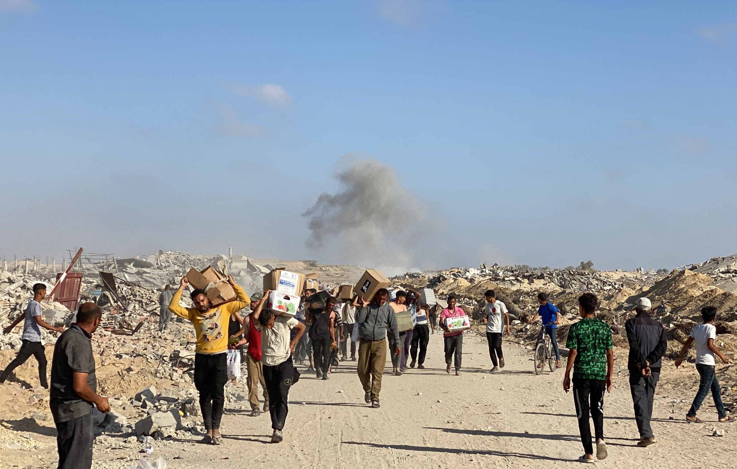 Des personnes marchent dans des décombres, portant des colis, avec de la fumée au loin.