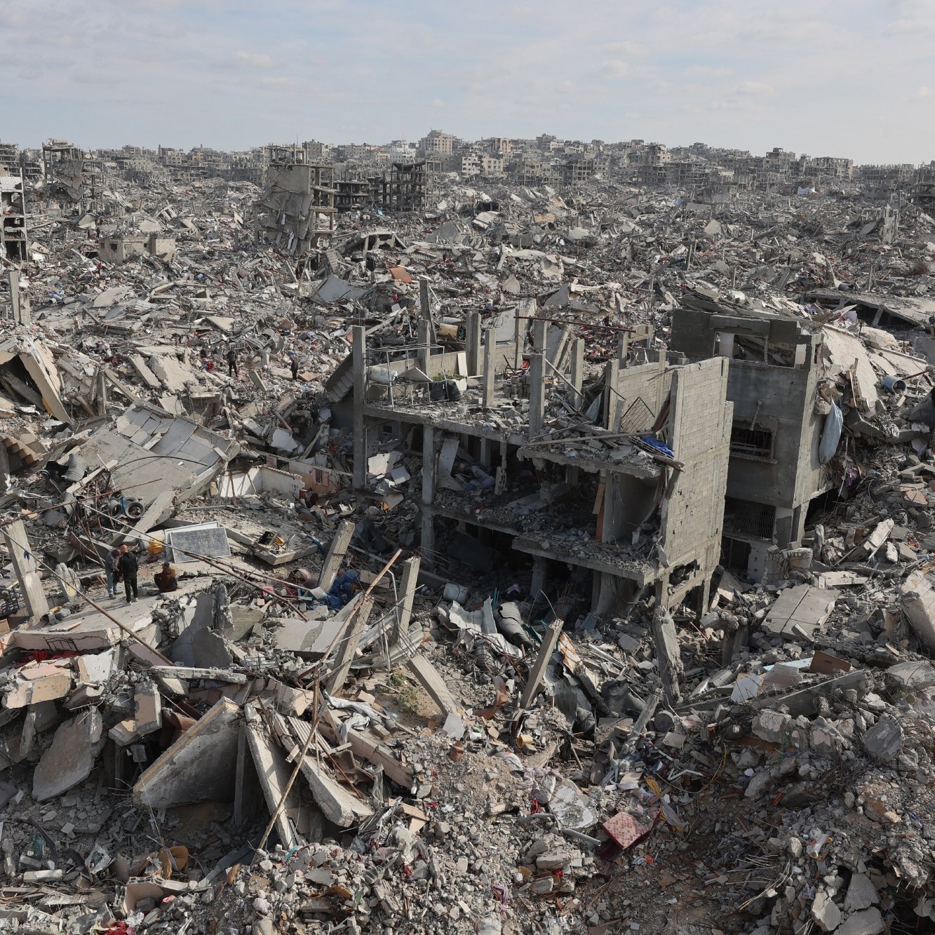 L'image montre un vaste paysage de débris et de bâtiments effondrés, engloutis dans les gravats. On aperçoit des structures en béton brisées et une atmosphère de destruction désolante.