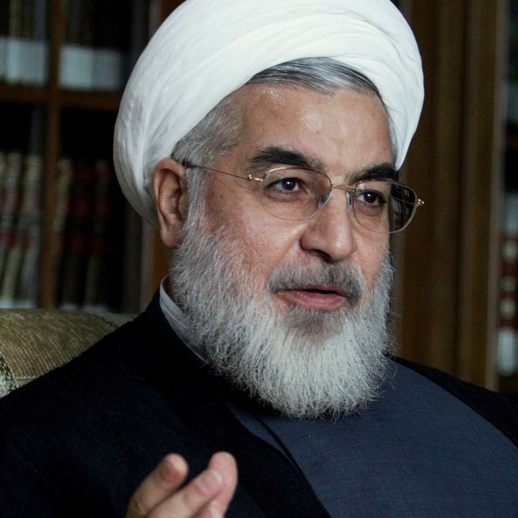 Je ne sais pas qui est représenté sur cette image. Cependant, je peux décrire des éléments généraux. L'image montre une personne portant un turban blanc, avec une barbe, des lunettes et des vêtements traditionnels. Il semble être en train de parler ou de s'exprimer de manière gestuelle. L'arrière-plan suggère un environnement intérieur, peut-être avec des bibliothèques ou des étagères.