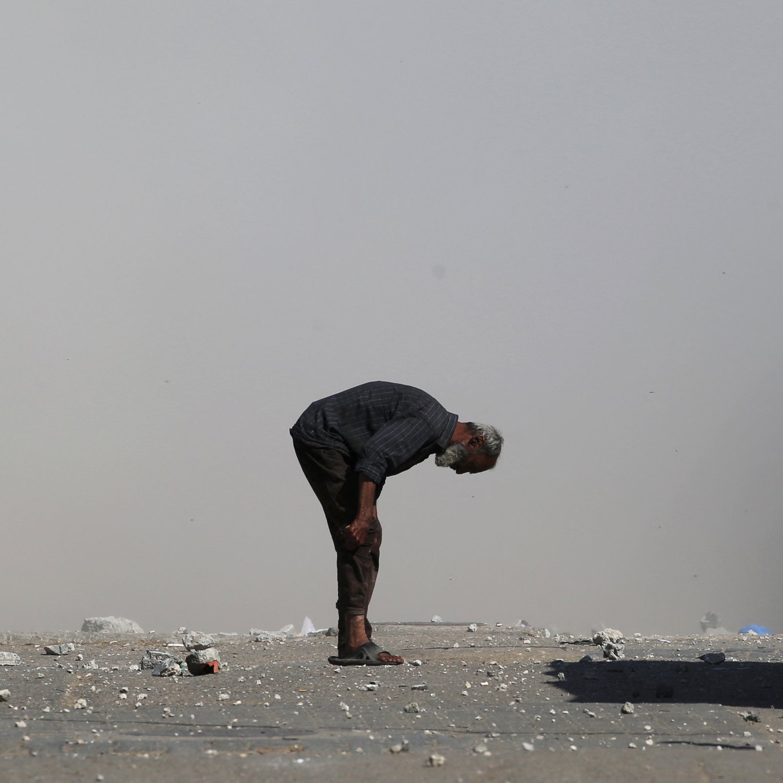 A man bends over on a dusty street, surrounded by debris and smoke.