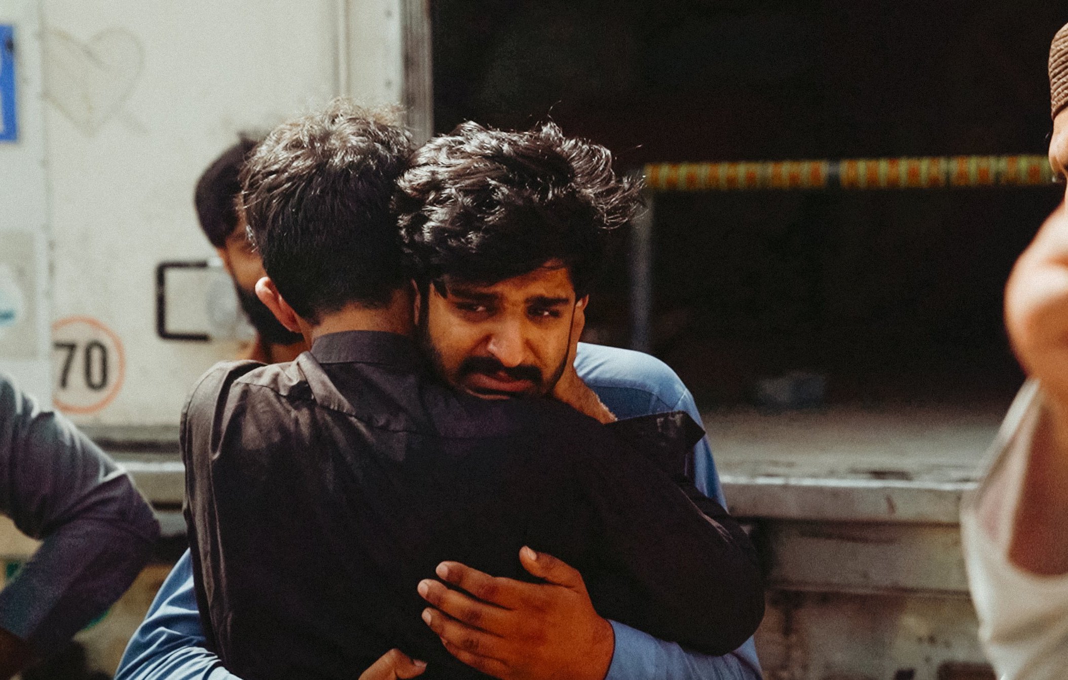 Two men embrace tightly, one looks visibly emotional, surrounded by others in a somber setting.