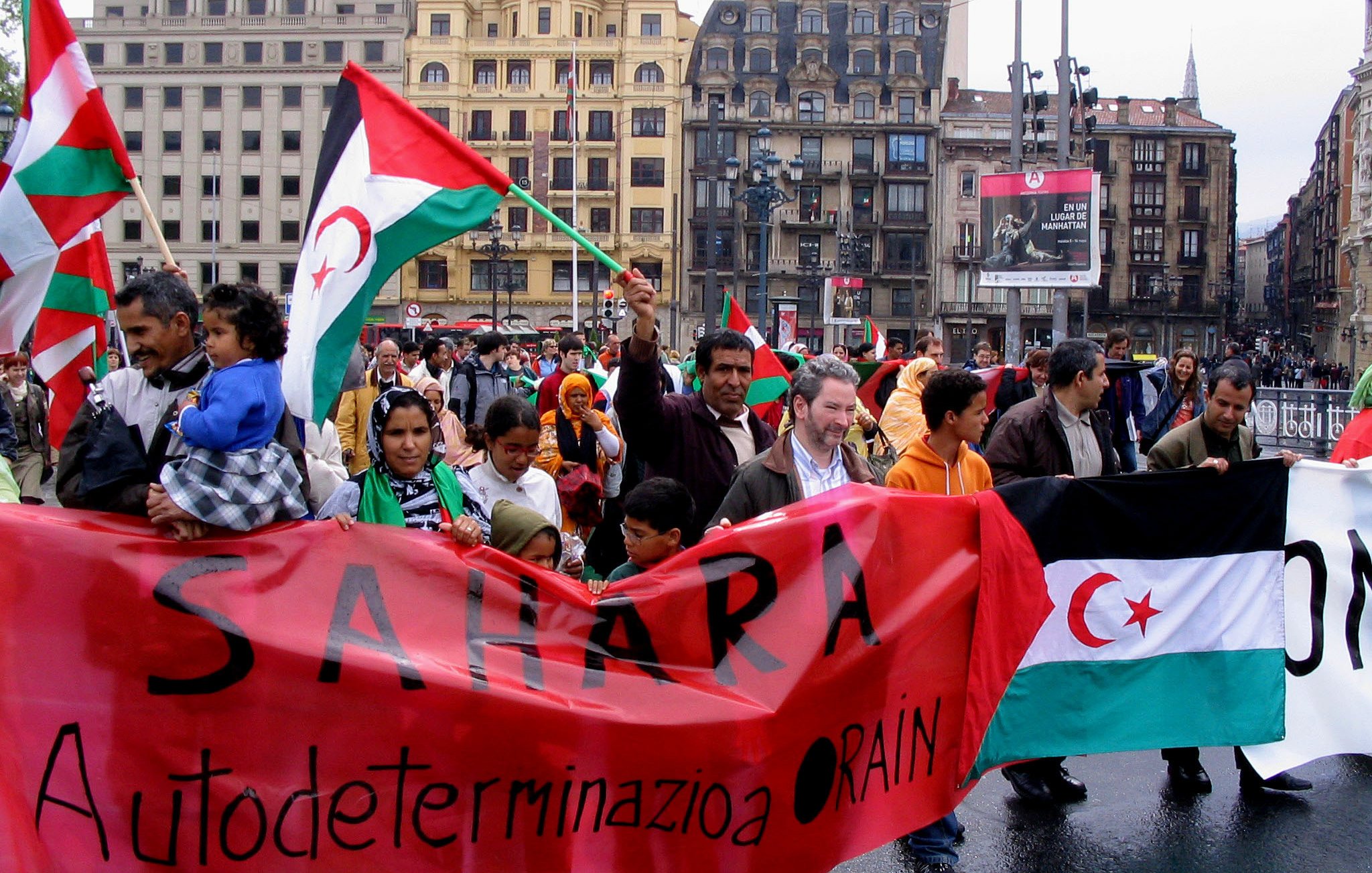 The image depicts a lively protest or demonstration taking place in an urban setting, likely in a city square. Participants are holding banners and flags, prominently featuring a design related to the Sahara. The banners include messages advocating for self-determination, with phrases like "Sahara Autodeterminazione" visible. The crowd consists of diverse individuals, including families and children, all appearing passionately engaged in the event. The background includes buildings and advertisements, adding to the urban atmosphere of the gathering. Rain can be seen drizzling, suggesting a potentially challenging weather environment for the demonstrators.