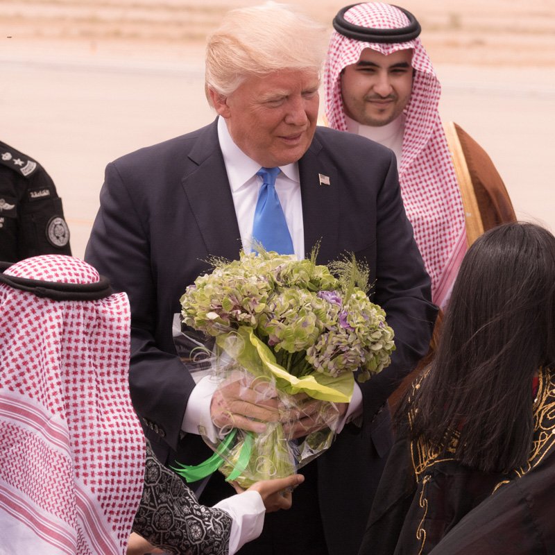 L'image montre un homme souriant, probablement un dirigeant, qui reçoit des fleurs à son arrivée. Il est entouré de plusieurs personnes portant des vêtements traditionnels, dont certains sont en tenue arabique. L'environnement semble être un aéroport, avec un paysage désertique en arrière-plan. Les personnes autour de lui semblent être accueillantes et amicales.